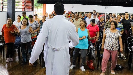 A imagem mostra uma atividade coletiva em um espaço interno, possivelmente uma unidade de saúde ou centro comunitário. Em primeiro plano, de costas para a câmera, está um profissional de saúde vestindo jaleco branco, conduzindo a atividade.  À frente dele, há um grupo numeroso de pessoas, em sua maioria adultos e idosos, participando ativamente. Muitos estão em pé, com braços e pernas em movimento, sugerindo uma prática de exercícios físicos orientados, como alongamento ou ginástica.  O ambiente tem piso de madeira e cadeiras ao fundo, indicando que o espaço também pode ser usado para reuniões ou grupos. A cena transmite energia, engajamento e promoção de saúde coletiva, com foco em atividade física e convivência.