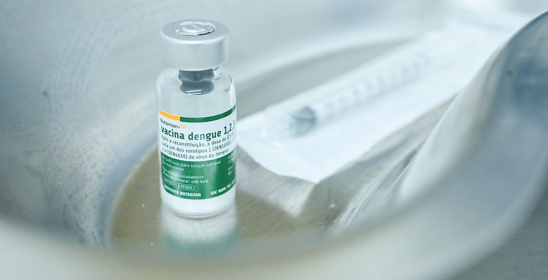 A imagem mostra um frasco de vacina posicionado dentro de uma bandeja metálica, com fundo levemente desfocado. O rótulo indica tratar-se de uma vacina contra a dengue. Ao lado do frasco, há uma seringa descartável ainda embalada, sugerindo preparo para aplicação.  O ambiente parece clínico, com iluminação suave e tons frios, transmitindo assepsia e cuidado. A composição destaca o frasco como elemento central, reforçando a ideia de prevenção e campanha de imunização.