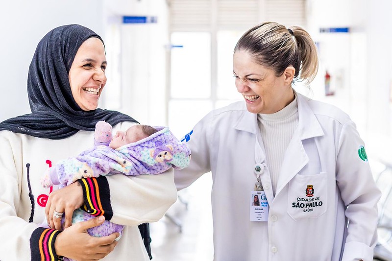 A imagem mostra um momento de cuidado e alegria em um ambiente de saúde. À esquerda, uma mulher usa um hijab escuro e segura um bebê recém-nascido envolto em um cobertor lilás com estampas delicadas. Ela sorri amplamente, olhando para a profissional ao seu lado.  À direita, uma profissional de saúde — vestindo jaleco branco com identificação da rede de saúde da cidade de São Paulo — também sorri, demonstrando empatia e acolhimento. Ela toca levemente o braço da mãe, em um gesto de proximidade e carinho.  O ambiente é claro, limpo e organizado, típico de uma unidade de saúde. A cena transmite sentimentos de acolhimento, confiança e cuidado, destacando o vínculo entre paciente, família e equipe de saúde, especialmente no contexto do nascimento e dos primeiros cuidados com o bebê.