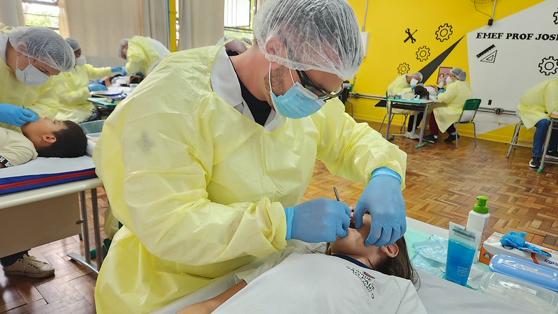 A imagem mostra escola municipal em que os alunos estão recebendo atendimento odontológico. Em primeiro plano, um profissional da saúde está realizando um procedimento odontológico em uma criança, que está em uma maca. Ele utiliza equipamentos de proteção individual completos: avental amarelo descartável, máscara cirúrgica, touca, luvas azuis e óculos de proteção. O profissional segura instrumentos odontológicos próximos à boca da paciente. A criança veste camiseta branca e permanece deitada enquanto recebe o atendimento. Ao lado da maca há materiais clínicos organizados, como frascos de álcool em gel, embalagens descartáveis e instrumentos odontológicos. Ao fundo, outras equipes também atendem crianças em macas semelhantes, indicando uma ação simultânea com vários atendimentos acontecendo no mesmo espaço. O ambiente possui piso de madeira e paredes amarelas decoradas com desenhos de engrenagens e elementos escolares.