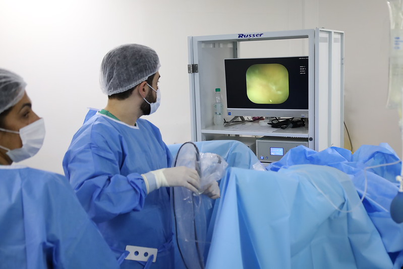 A imagem mostra um procedimento médico realizado em ambiente hospitalar ou centro cirúrgico. Em primeiro plano, aparecem profissionais de saúde usando aventais cirúrgicos azuis, máscaras, luvas e toucas descartáveis, seguindo protocolos de assepsia. Um dos profissionais segura um equipamento flexível semelhante a um endoscópio enquanto observa atentamente um monitor. Na tela é exibida uma imagem interna do corpo humano captada por vídeo, indicando que se trata de um procedimento minimamente invasivo, como uma endoscopia, colonoscopia, broncoscopia ou outro exame/intervenção guiado por câmera. O paciente está coberto por campos cirúrgicos azuis estéreis, que ocupam boa parte da cena. O ambiente é limpo, bem iluminado e equipado com tecnologia médica. A fotografia transmite uma atmosfera de cuidado especializado, precisão técnica e trabalho em equipe, destacando o uso de recursos diagnósticos e terapêuticos modernos na assistência à saúde.