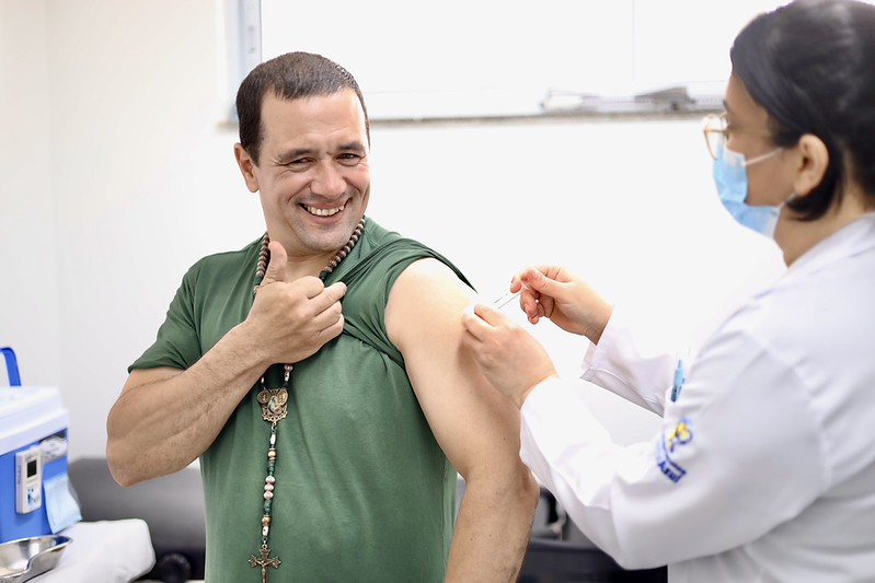 A imagem mostra uma cena de vacinação em um ambiente de saúde.  Um homem adulto, vestindo uma camiseta verde e um colar com contas e um crucifixo, sorri enquanto recebe uma injeção no braço esquerdo. Com uma das mãos, ele segura a manga da camiseta levantada para facilitar a aplicação. Sua expressão é descontraída e positiva, transmitindo confiança e tranquilidade durante o procedimento.  À direita da imagem, uma profissional de saúde, vestida com jaleco branco e máscara cirúrgica azul, prepara ou aplica a vacina. Ela aparece de perfil, concentrada no atendimento. O ambiente é limpo, bem iluminado e tem características típicas de um consultório ou sala de vacinação, com equipamentos médicos e superfícies clínicas ao fundo.