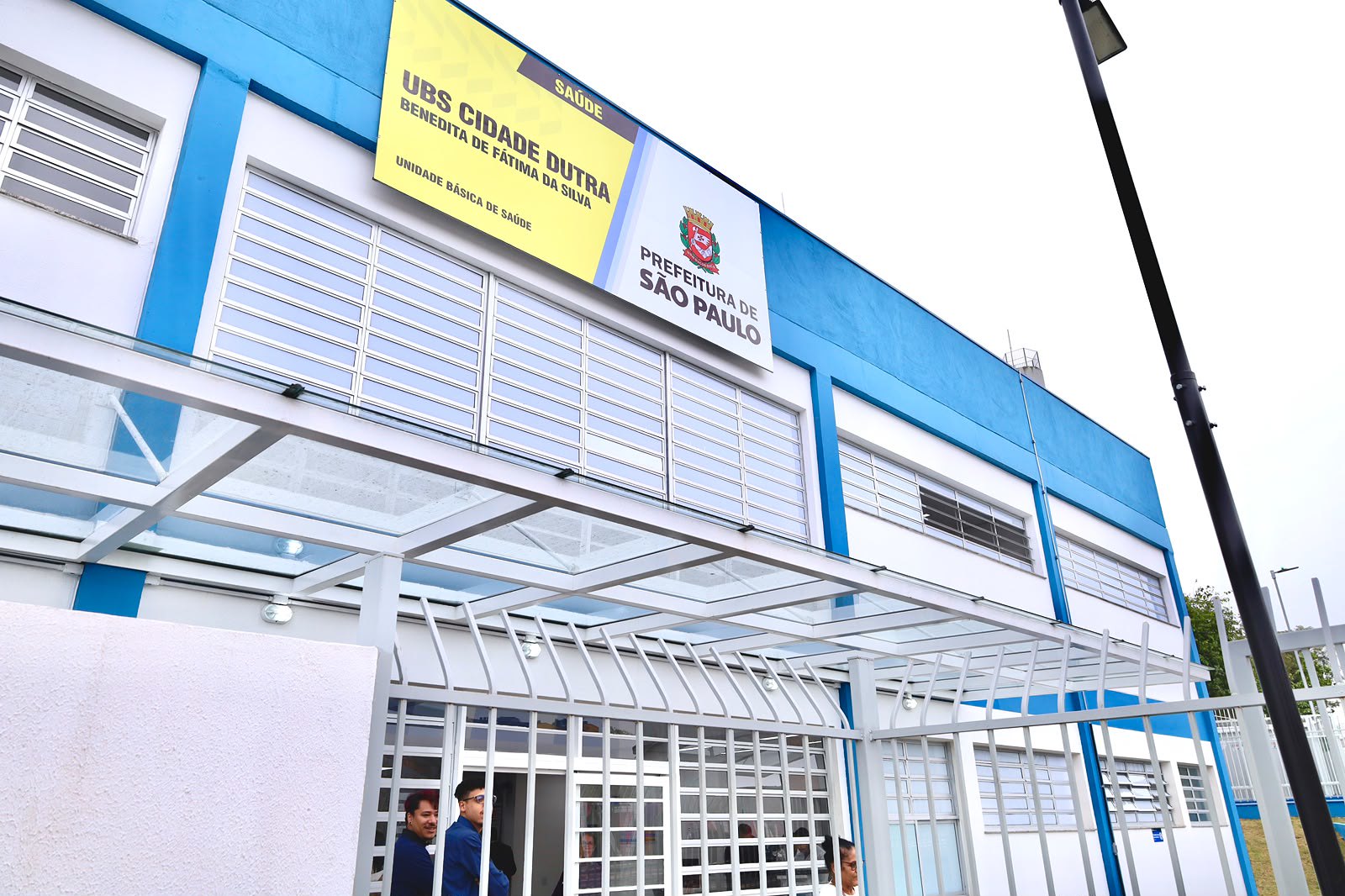 achada da UBS Cidade Dutra – Benedita de Fátima da Silva, prédio de dois pavimentos nas cores branca e azul. Na parte superior, uma placa de identificação da unidade ao lado do brasão da Prefeitura de São Paulo. A entrada é protegida por uma cobertura de vidro e cercada por gradil metálico, enquanto algumas pessoas entram na unidade. A imagem destaca a nova estrutura da Unidade Básica de Saúde.