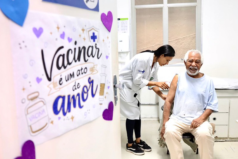 Profissional de saúde aplica vacina no braço de um homem idoso, que sorri sentado em uma cadeira durante o atendimento. Ao lado, um cartaz colorido destaca a mensagem “Vacinar é um ato de amor”.