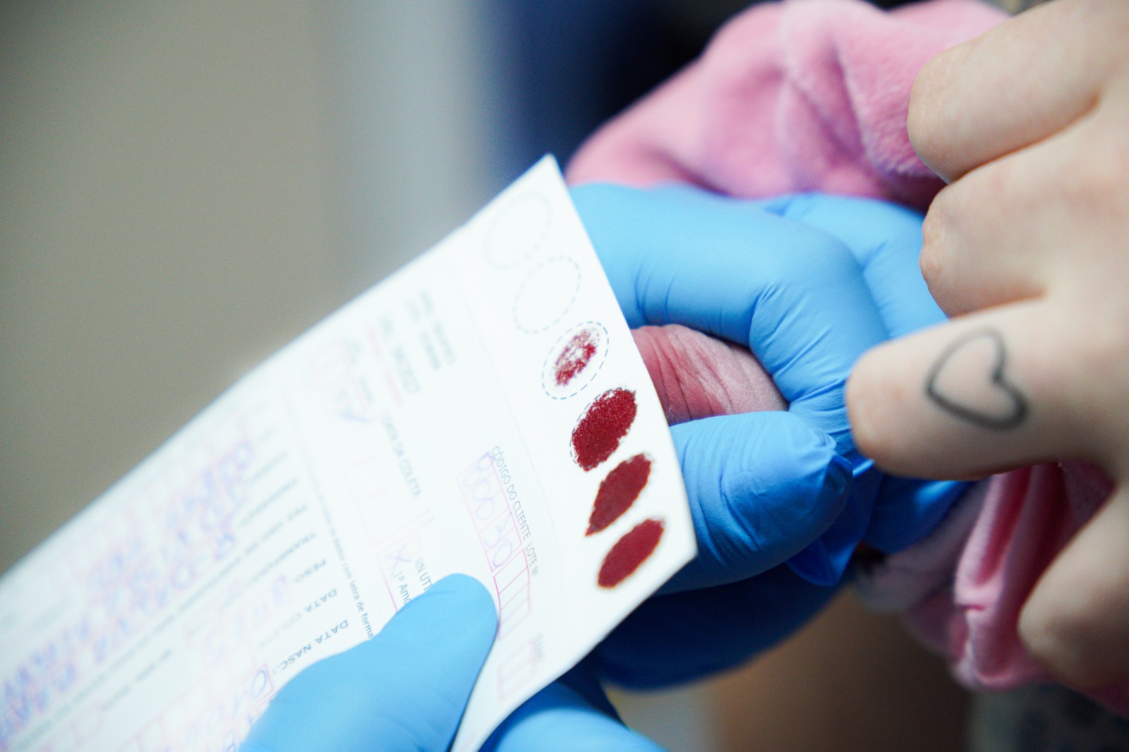 A imagem mostra um close-up de um procedimento de coleta de sangue por punção digital para realização de teste rápido ou exame laboratorial. Em destaque, uma profissional utilizando luvas descartáveis azuis segura cuidadosamente o dedo de uma pessoa enquanto coleta pequenas gotas de sangue em um cartão ou formulário específico para análise. Sobre o cartão, é possível observar várias gotas de sangue depositadas em áreas circulares demarcadas para a amostra. A pessoa atendida veste uma peça de roupa rosa e tem uma pequena tatuagem em formato de coração em um dos dedos visíveis. O enquadramento aproxima a cena dos detalhes das mãos e do material de coleta, enquanto o fundo aparece desfocado, direcionando a atenção para o procedimento. A fotografia transmite um ambiente de cuidado e atendimento em saúde, ilustrando uma etapa comum de testagem rápida ou exames que utilizam amostras de sangue obtidas por punção na ponta do dedo.