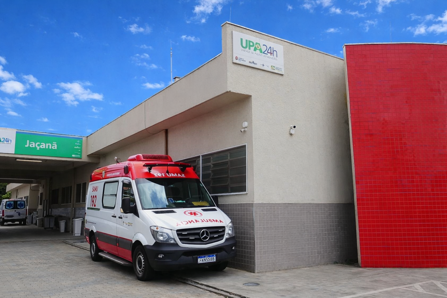 Entrada da UPA 24h Jaçanã, com ambulância do SAMU posicionada para atendimento. A imagem destaca a estrutura da unidade, com fachada clara, parede vermelha e céu azul, reforçando o cuidado permanente com a população.