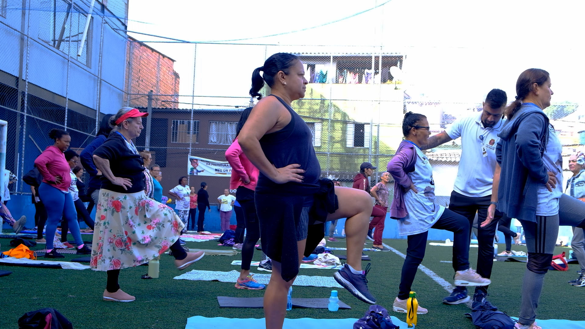 A imagem mostra um grupo de pessoas participando do Pilates na Quebrada em um campo de futebol ao ar livre. A turma é composta principalmente por mulheres, de diferentes idades, e todas estão fazendo um exercício que envolve levantar uma perna e manter uma das mãos na cintura para equilíbrio. O professor Pedro, vestido de branco, orienta os participantes. Ao fundo, é possível ver um mural com a logomarca da prefeitura e algumas construções residenciais.