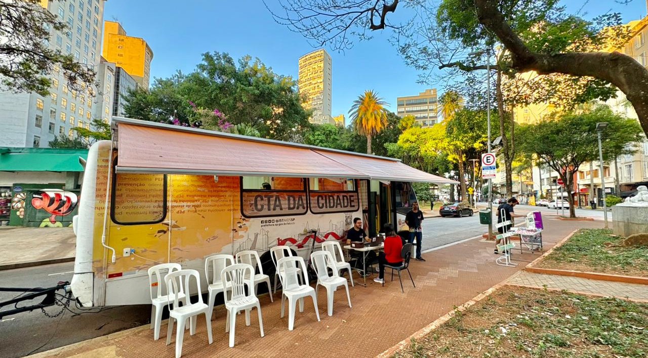 A imagem mostra uma unidade móvel de atendimento instalada em uma praça ou área pública arborizada de uma região central da cidade. &nbsp;Em destaque está um trailer adaptado, identificado como “CTA da Cidade”, sigla normalmente associada aos Centros de Testagem e Aconselhamento em saúde. O veículo possui uma cobertura retrátil aberta sobre a calçada, criando uma área de atendimento ao público. Em frente ao trailer há várias cadeiras plásticas brancas organizadas para espera, enquanto algumas pessoas conversam ou realizam atendimentos em mesas instaladas ao lado da unidade. &nbsp;O cenário urbano é composto por prédios altos ao fundo, árvores de grande porte, palmeiras e uma praça com áreas de circulação de pedestres. A luz do sol ilumina a cena, sugerindo que a atividade ocorre durante o dia.