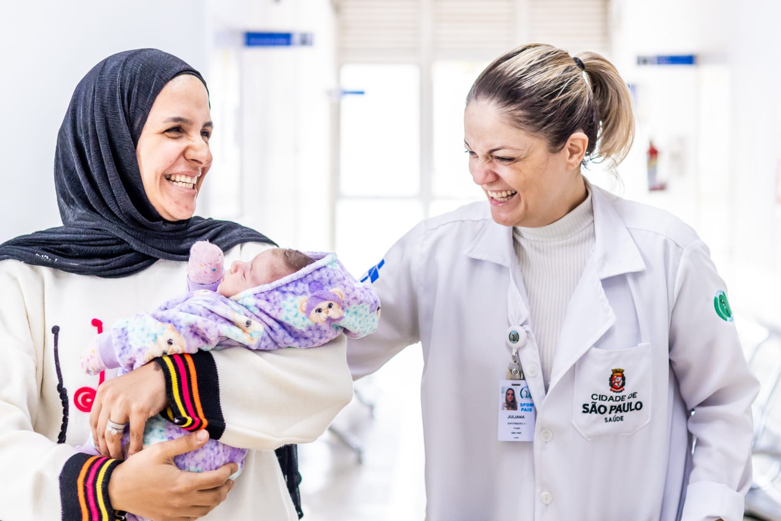 A imagem mostra uma cena na AMA/UBS Pari. À esquerda, há uma mulher sorridente usando um lenço (hijab) na cabeça. Ela está segurando um bebê recém-nascido envolto em um cobertor roxo com estampas delicadas. O bebê parece estar dormindo tranquilamente. À direita, há uma profissional de saúde vestindo um jaleco branco com identificação da área de saúde da cidade de São Paulo. Ela também está sorrindo e olhando com carinho para o bebê, com uma das mãos apoiada de forma acolhedora no ombro da mãe. O ambiente ao fundo é claro, limpo e organizado, com aparência típica de um espaço hospitalar. A cena transmite um clima de alegria, acolhimento e cuidado. 