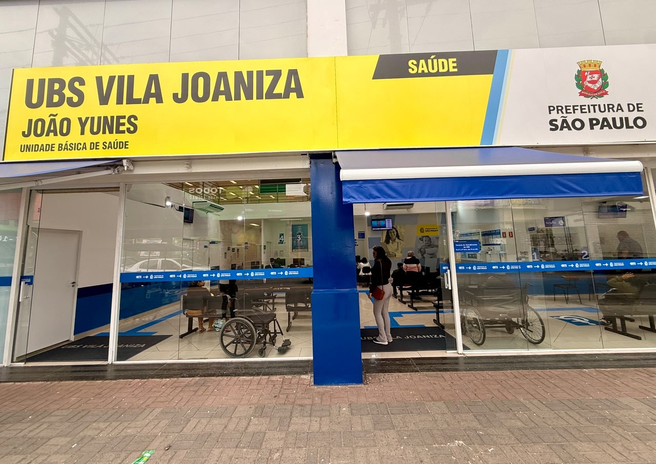 A imagem mostra a fachada da Unidade Básica de Saúde (UBS) Vila Joaniza – João Yunes, localizada no município de São Paulo. Na parte superior da fachada, destaca-se uma grande placa nas cores amarela, preta, azul e branca, com os dizeres “UBS Vila Joaniza – João Yunes – Unidade Básica de Saúde”. À direita, aparecem o brasão da cidade e a identificação da Prefeitura de São Paulo, além da palavra “Saúde”. A entrada da unidade é composta por amplas portas e paredes de vidro, permitindo visualizar o interior do prédio. Dentro da recepção, observam-se cadeiras para espera, algumas pessoas sentadas e outras circulando pelo local. Há também cadeiras de rodas posicionadas próximas à entrada, indicando acessibilidade para usuários com mobilidade reduzida. A fachada possui uma coluna central azul e uma cobertura retrátil azul sobre a entrada principal. O piso externo é revestido com blocos de concreto intertravados, formando uma calçada ampla em frente à unidade. A imagem transmite a aparência de uma unidade de saúde pública organizada, moderna e acessível, com ambiente interno iluminado e sinalização institucional bem visível.