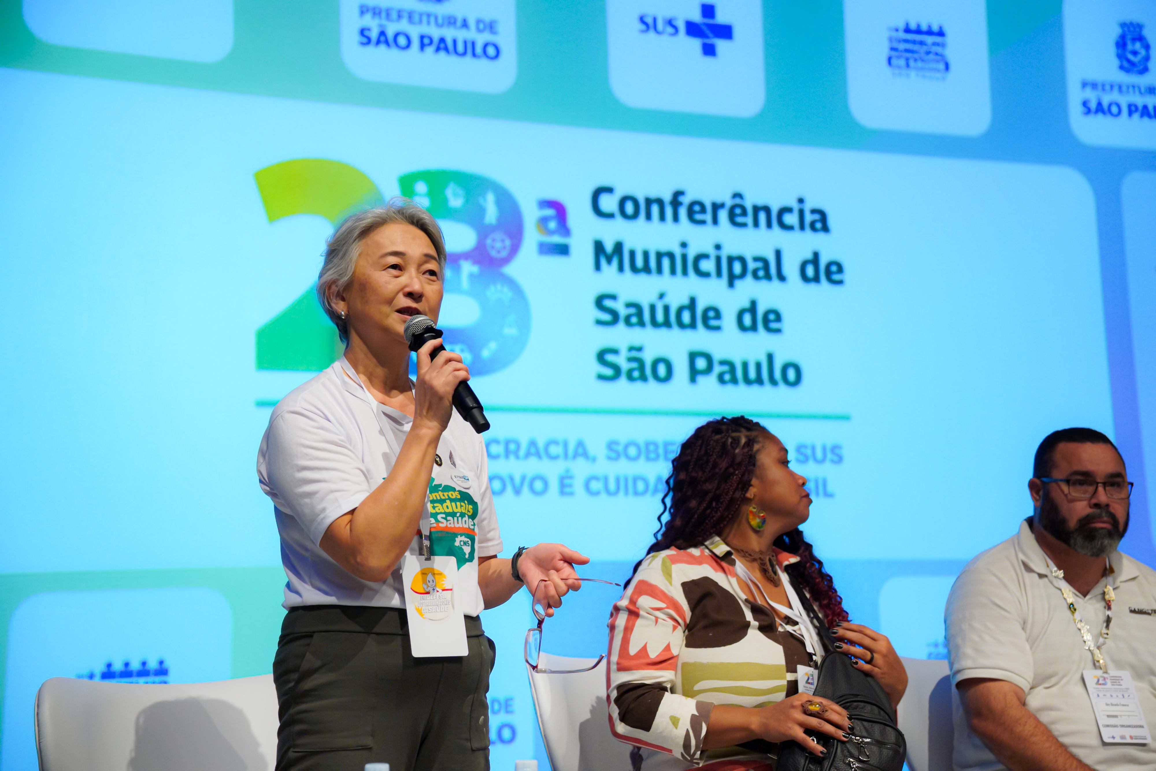 A foto mostra um momento de fala durante a 23ª Conferência Municipal de Saúde de São Paulo, realizada em um auditório. Em primeiro plano, uma mulher de cabelos grisalhos curtos está em pé, segurando um microfone e falando ao público. Ela veste uma camiseta branca relacionada ao evento, calça verde-escura e um crachá de identificação preso ao pescoçoAo lado dela, sentados em poltronas brancas sobre o palco, estão outros dois participantes do painel. Ao centro, uma mulher de cabelos longos e trançados usa uma blusa estampada em tons de branco, marrom, verde e coral. À direita, um homem de barba, óculos e camiseta polo branca também está sentado, usando um crachá do evento e observando a apresentação. Ao fundo, um grande telão exibe a identidade visual da conferência, com o número 23 em destaque e o texto