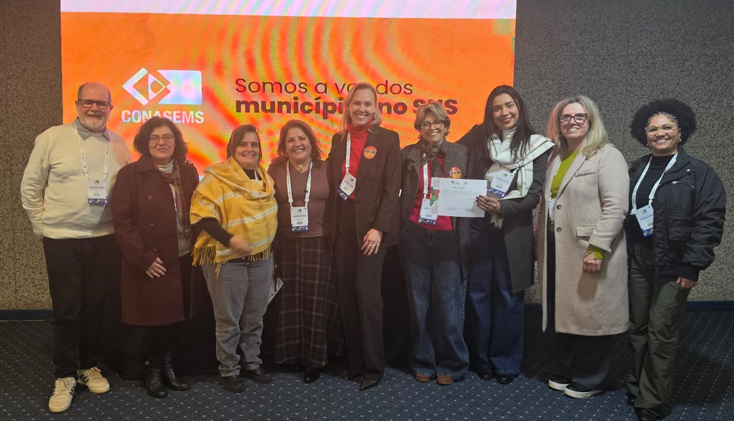 A imagem mostra um grupo de nove pessoas posando lado a lado em um ambiente interno, durante o 39° Conasems. Todas estão de frente para a câmera, sorrindo, e usam crachás de identificação presos ao pescoço, característicos de congressos ou encontros profissionais. Ao fundo, há um grande painel eletrônico na cor laranja com o logotipo do Conasems e a frase: