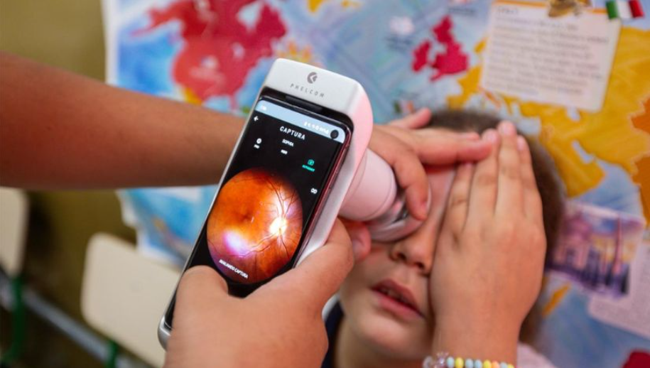A imagem mostra uma criança realizando um exame ocular utilizando um aparelho. Uma pessoa segura o equipamento, que está capturando uma imagem do fundo do olho da criança. Na tela do dispositivo, é possível ver a retina com detalhes, indicando que o exame está em andamento. A criança cobre um dos olhos com a mão enquanto o outro está sendo examinado. Ao fundo, há um mapa mundi colorido.