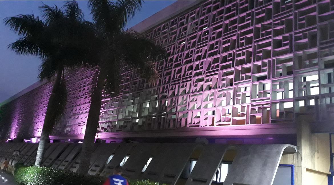 A imagem mostra a fachada iluminada do Hospital Municipal Cidade Tiradentes – Carmen Prudente, localizado na zona leste de São Paulo. A arquitetura moderna é marcada por uma grande parede com elementos vazados em formas geométricas retangulares, criando um efeito visual dinâmico. A iluminação em tom roxo ou lilás valoriza a estrutura, destacando os relevos da fachada durante a noite. Em primeiro plano, há palmeiras e vegetação aparada, além de pessoas circulando próximo ao prédio. A cena transmite um ambiente urbano bem cuidado e acolhedor, com iluminação que pode estar relacionada a alguma campanha de saúde ou conscientização.
