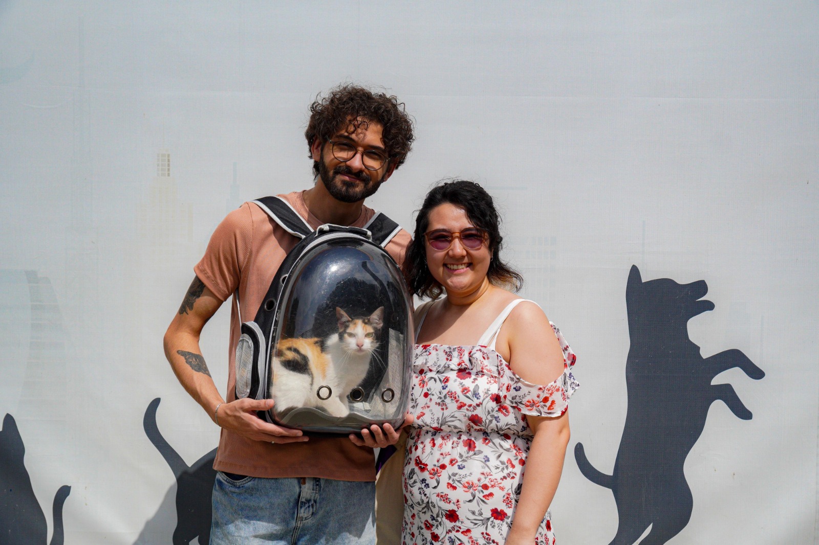 A foto mostra duas pessoas sorrindo para a câmera, lado a lado, em frente a um fundo claro decorado com grandes silhuetas de gatos. À esquerda, há um homem de cabelos cacheados, barba e óculos, usando uma camiseta em tom terracota. Ele segura uma mochila/caixa de transporte transparente para gatos. Dentro da caixa está um gato tricolor (branco, laranja e preto), olhando diretamente para a câmera. À direita, há uma mulher de cabelos escuros, usando óculos de sol com lentes avermelhadas e um vestido branco estampado com flores vermelhas e cinzas. A iluminação é forte, sugerindo um ambiente externo em um dia de sol.