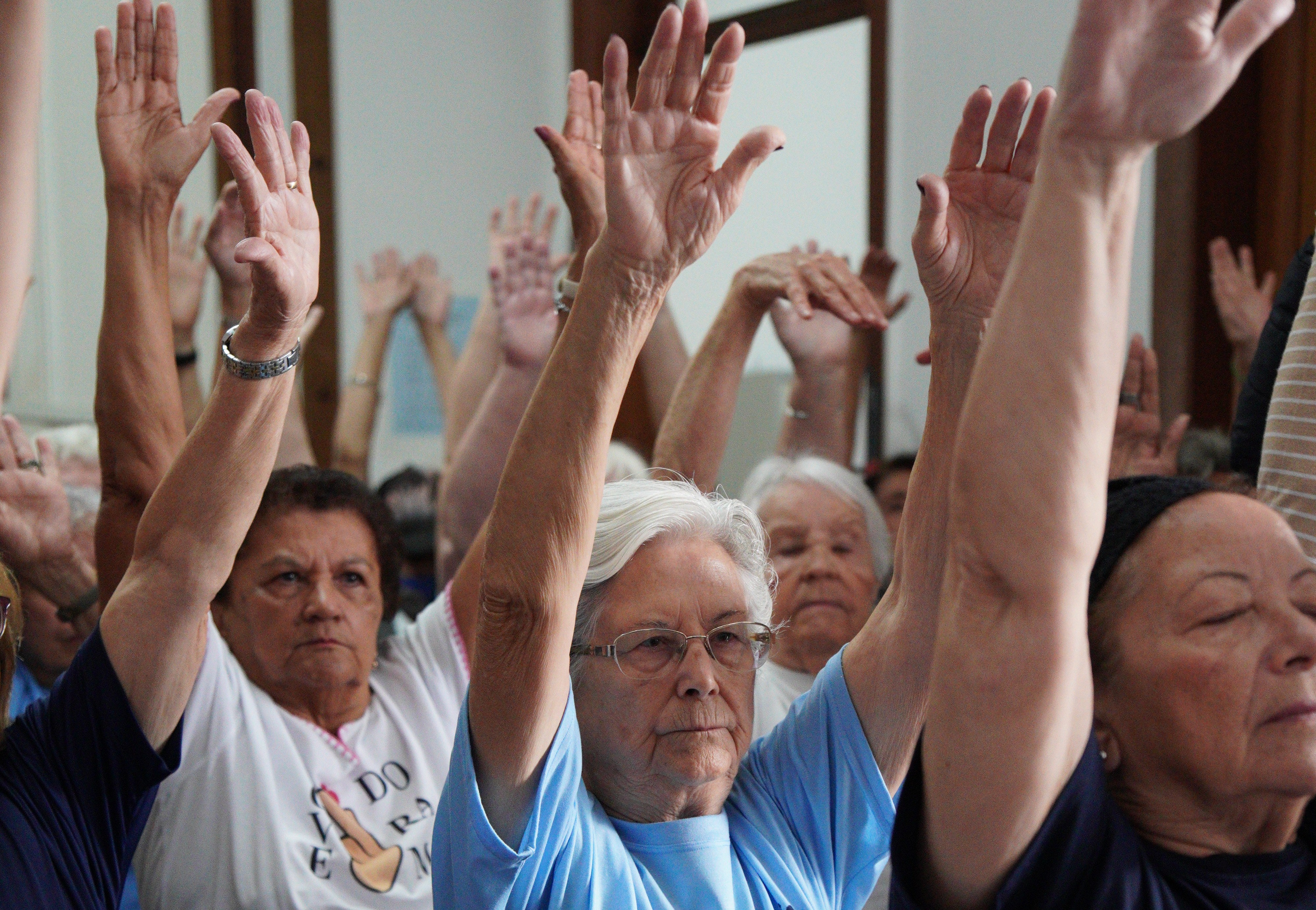 A imagem apresenta um grupo de mulheres idosas em uma atividade coletiva em ambiente fechado. Elas aparecem em primeiro plano e ao fundo, com os braços erguidos e as palmas das mãos voltadas para a frente, sugerindo um momento de exercício físico, alongamento ou uma dinâmica de grupo.  No centro da composição, destaca-se uma senhora de cabelos brancos e óculos, vestindo uma camiseta azul clara, com uma expressão serena e concentrada. Ao lado dela, outras mulheres participam do movimento, vestindo roupas confortáveis, como camisetas brancas e escuras. A iluminação é suave, e o foco da fotografia recai principalmente sobre as figuras centrais, criando uma sensação de engajamento e vitalidade.