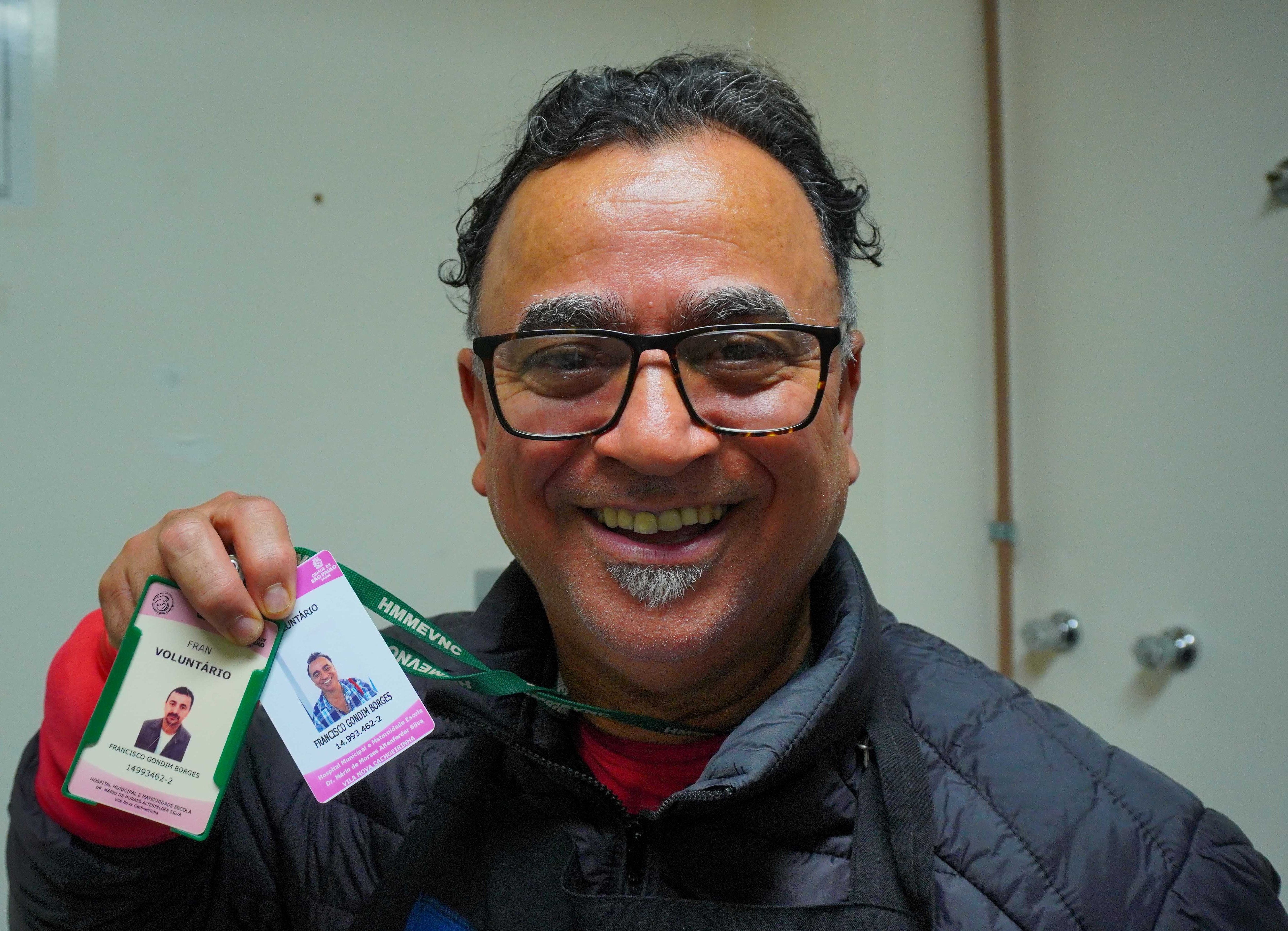 Retrato em close de Francisco, um homem sorridente, em ambiente interno de hospital. Ele tem cabelo preto curto com alguns fios grisalhos e levemente ondulados, entradas discretas, sobrancelhas espessas e um pequeno cavanhaque grisalho no queixo. Usa óculos de armação retangular escura. Veste blusa vermelha por baixo de uma jaqueta preta acolchoada e está com um avental escuro.  Com a mão esquerda, ele ergue dois crachás presos a um cordão verde. O crachá da esquerda, com moldura verde e a palavra “VOLUNTÁRIO” visível, foi o primeiro que ele recebeu ao iniciar o voluntariado no hospital. À direita, há um segundo crachá, com moldura lilás/rosa, também com sua foto. A expressão aberta e o gesto com os crachás transmitem orgulho e alegria.