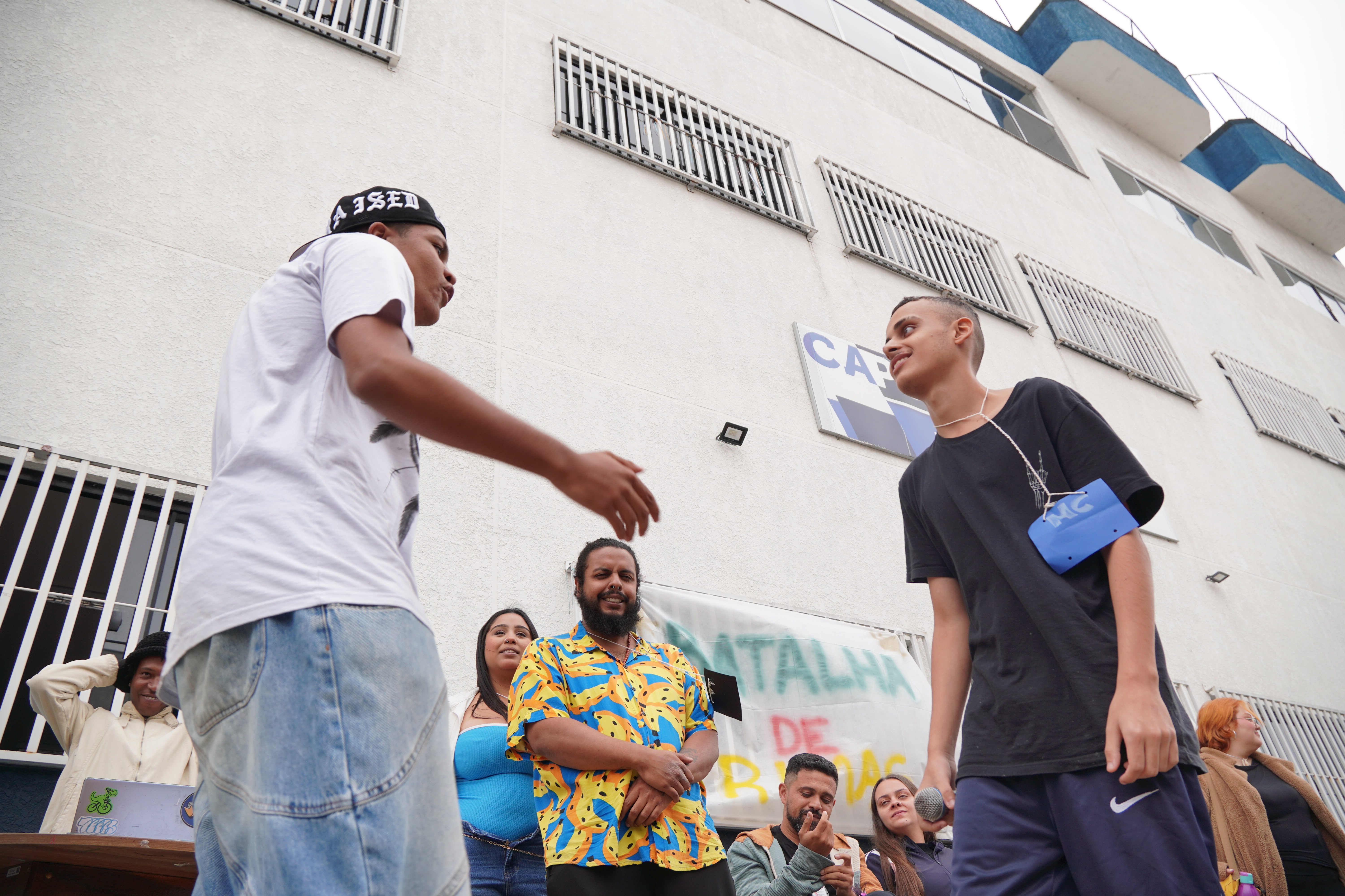 A imagem registra dois jovens negros se confrontando em uma batalha de rimas ao ar livre. O jovem da direita segura um microfone, e o da esquerda gesticula em direção ao outro. O público assiste em frente a um banner de evento e a um prédio grande de cor clara, com a cena capturada em uma perspectiva de baixo para cima.