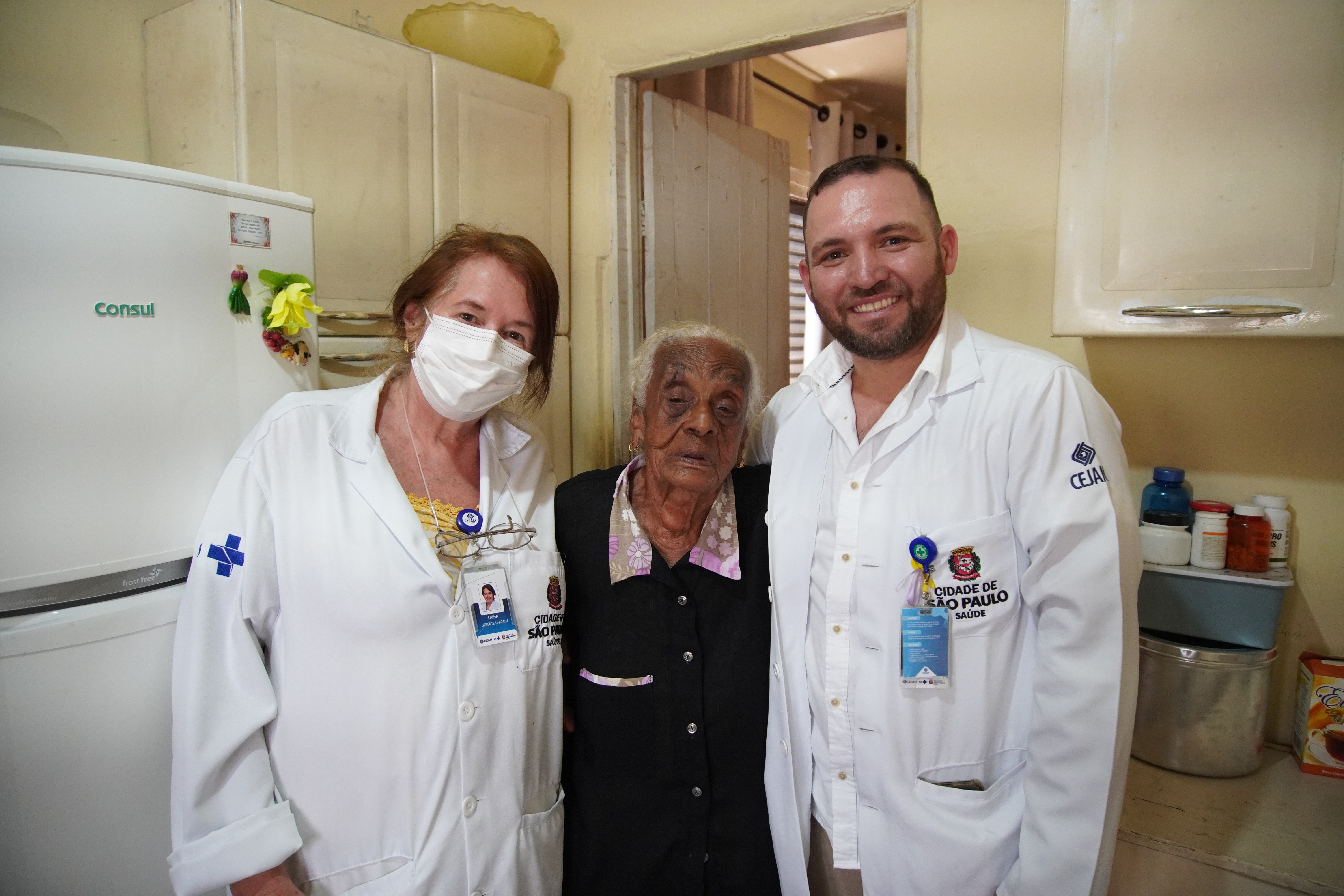 A imagem mostra três pessoas em um ambiente interno, possivelmente uma cozinha. No centro está uma senhora idosa de pele escura, cabelos brancos e curtos, vestindo uma camisa escura com detalhes florais. Ela está ladeada por dois profissionais de saúde que usam jalecos brancos com identificações visíveis.  À esquerda da imagem está uma mulher com cabelos castanhos claros e soltos, parcialmente cobertos por uma máscara branca. Ela usa um jaleco com o brasão da Cidade de São Paulo – Saúde e um crachá com sua foto.  À direita da senhora idosa, está um homem sorridente, de barba aparada, também vestindo jaleco com o brasão da Cidade de São Paulo – Saúde e o logotipo do CEJAM (Centro de Estudos e Pesquisas “Dr. João Amorim”). Ele também usa um crachá de identificação.  Ao fundo, é possível ver uma geladeira, armários, utensílios e frascos de medicamentos, sugerindo que o atendimento ocorre na residência da idosa. A imagem transmite um momento de cuidado e acolhimento, provavelmente de um atendimento domiciliar da rede pública de saúde.