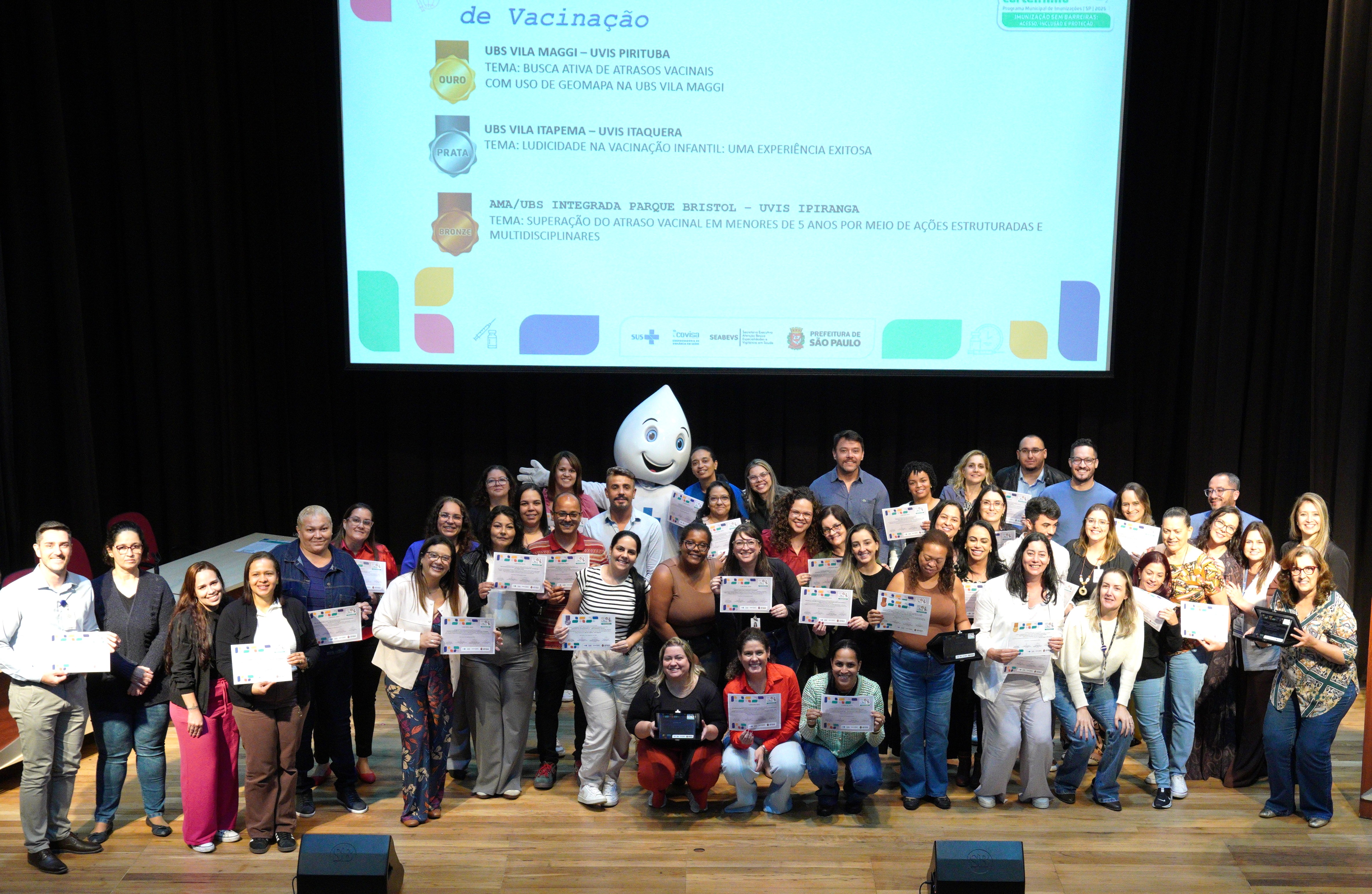 Foto de grupo em um palco de auditório. Cerca de quarenta pessoas adultas, de diferentes gêneros, idades e tons de pele, estão reunidas em três fileiras. A maioria sorri e segura certificados impressos com as duas mãos, voltados para a câmera. As pessoas vestem roupas variadas, em tons claros e escuros, incluindo camisas, blusas, calças e vestidos, em estilo casual a social.  No centro do grupo, há um personagem mascote em formato de gota d’água branca, com olhos grandes e sorriso, simbolizando saúde ou vacinação. Algumas pessoas estão em pé e outras agachadas na primeira fileira, criando uma composição organizada para a foto.  Ao fundo, acima do grupo, há um telão de projeção com um slide institucional. O slide apresenta o tema relacionado à vacinação e traz menções a unidades de saúde (UBS e AMA/UBS), além de categorias de premiação representadas por medalhas de ouro, prata e bronze. Na parte inferior do slide, aparecem logotipos institucionais, incluindo o da Prefeitura de São Paulo e do SUS.  O ambiente é bem iluminado, com cortinas escuras atrás do telão e piso de madeira no palco, sugerindo um evento formal de reconhecimento ou premiação na área da saúde pública.