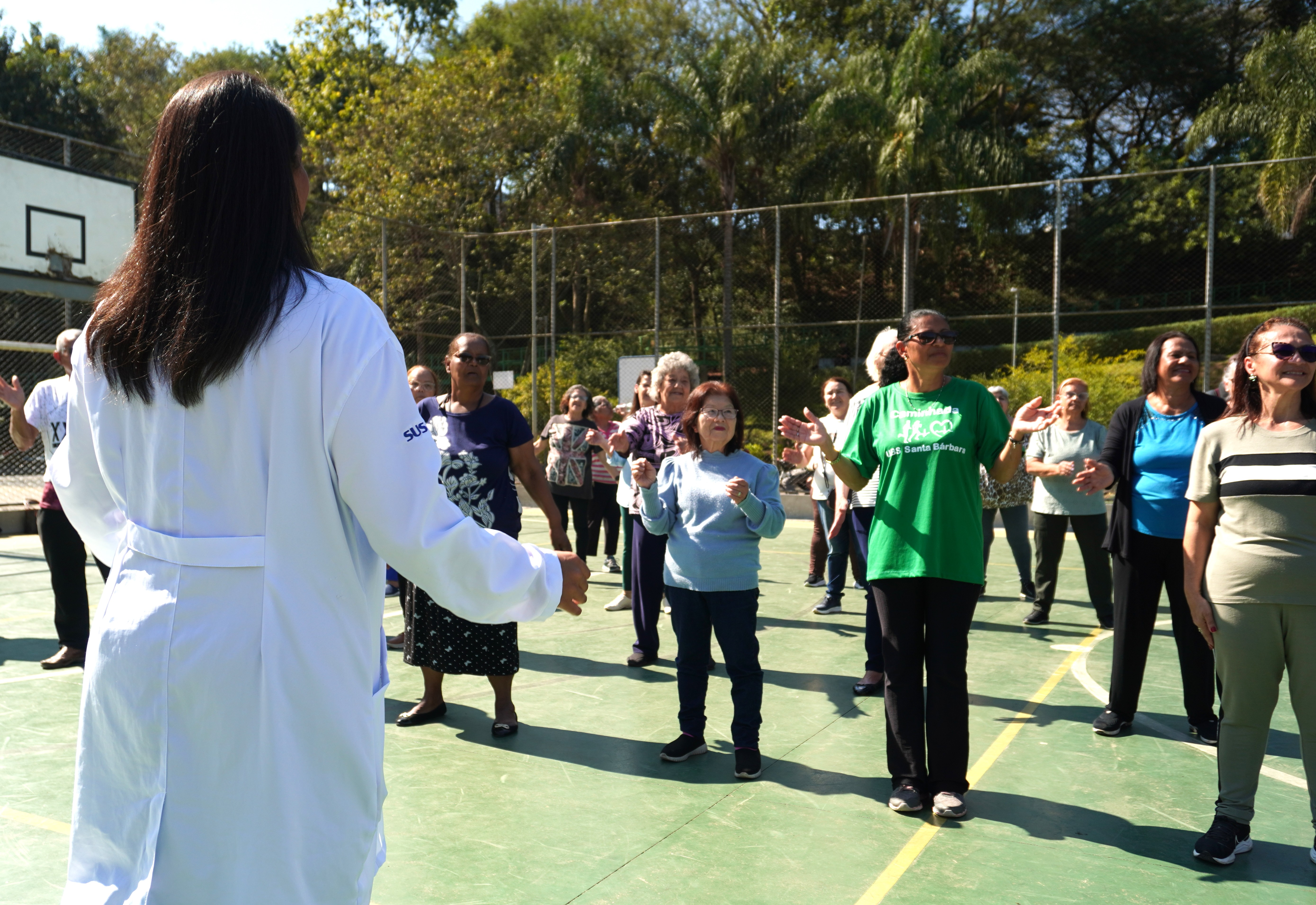 A imagem mostra uma atividade coletiva ao ar livre, em uma quadra esportiva cercada por árvores.  Em primeiro plano, de costas para a câmera, há uma profissional da saúde vestindo um jaleco branco com o logotipo do SUS, aparentemente orientando ou conduzindo a atividade. Ela estende os braços, como se estivesse demonstrando movimentos.  À frente dela, um grupo majoritariamente formado por mulheres, muitas delas idosas, acompanha os gestos. As participantes estão em pé, com os braços erguidos, sugerindo que realizam exercícios leves, alongamentos ou uma prática corporal orientada.  O ambiente é ensolarado e transmite sensação de bem-estar. Algumas mulheres usam roupas confortáveis para atividade física, e uma delas veste uma camiseta verde com referência a uma unidade de saúde (“UBS Santa Bárbara”).  A cena indica uma ação de promoção da saúde, provavelmente ligada à atenção básica, com foco em atividade física, convivência e qualidade de vida na comunidade.