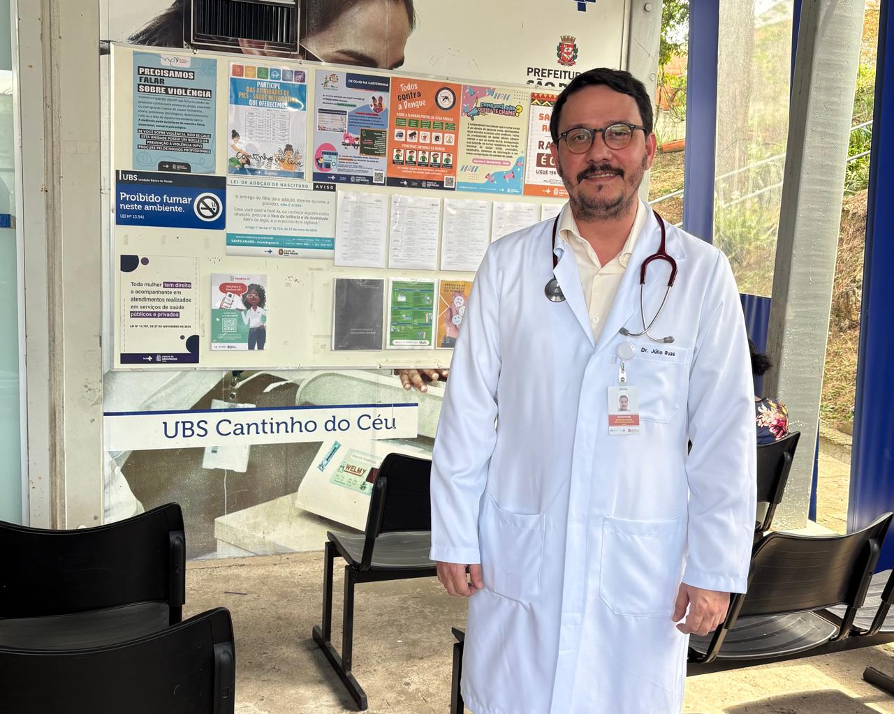 A imagem mostra um profissional de saúde de pé, vestindo jaleco branco e estetoscópio no pescoço. Ele está sorridente e usa óculos. No crachá preso ao jaleco, é possível ler “Dr. Júlio Ruas”.  Ao fundo, há um painel com cartazes informativos e comunicados sobre saúde pública, fixado na parede de uma unidade identificada como UBS Cantinho do Céu, administrada pela Prefeitura de São Paulo. O ambiente parece ser a área de espera da unidade, com cadeiras pretas dispostas no local. A cena transmite um clima de atendimento humanizado e de atuação médica em uma unidade básica de saúde da rede municipal.