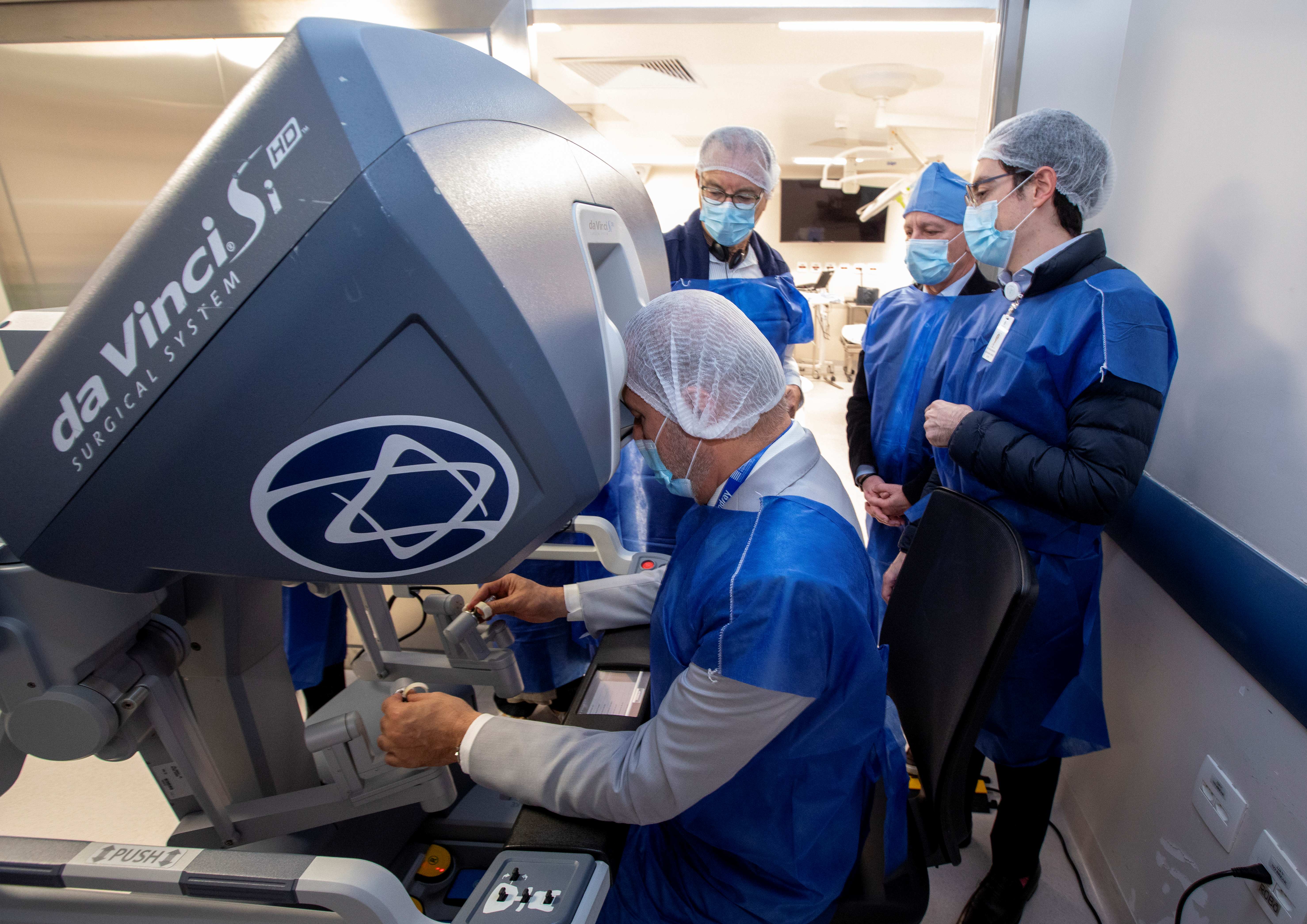 A imagem mostra uma cirurgia robótica em um ambiente hospitalar ou de centro cirúrgico. Quatro pessoas estão utilizando roupas e equipamentos de proteção: aventais azuis descartáveis, toucas e máscaras. No primeiro plano, uma pessoa está sentada e operando um equipamento grande e moderno, que é um sistema de cirurgia robótica. Essa pessoa olha para dentro do visor da máquina e manuseia dois controles com as mãos, como se estivesse treinando ou realizando um procedimento por meio do robô. Ao redor dela, outras três pessoas observam atentamente o processo.