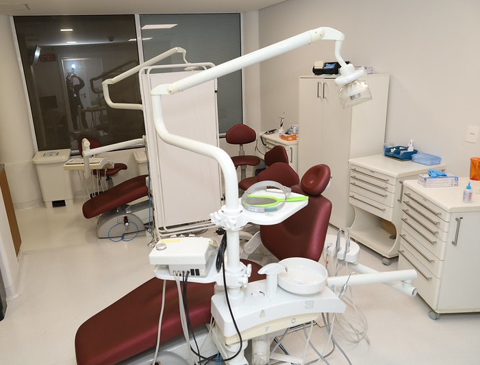 A imagem mostra um consultório odontológico moderno e bem iluminado. No centro da sala há uma cadeira odontológica reclinável na cor vinho, acompanhada de diversos equipamentos acoplados, como refletor de luz, bandejas de instrumentos e mangueiras para procedimentos dentários. Ao lado da cadeira principal há um banco giratório para o profissional e armários brancos com gavetas , onde ficam armazenados materiais e equipamentos clínicos. Sobre as bancadas aparecem alguns itens descartáveis e utensílios odontológicos. O ambiente possui piso claro e paredes em tons neutros, transmitindo sensação de limpeza e organização.