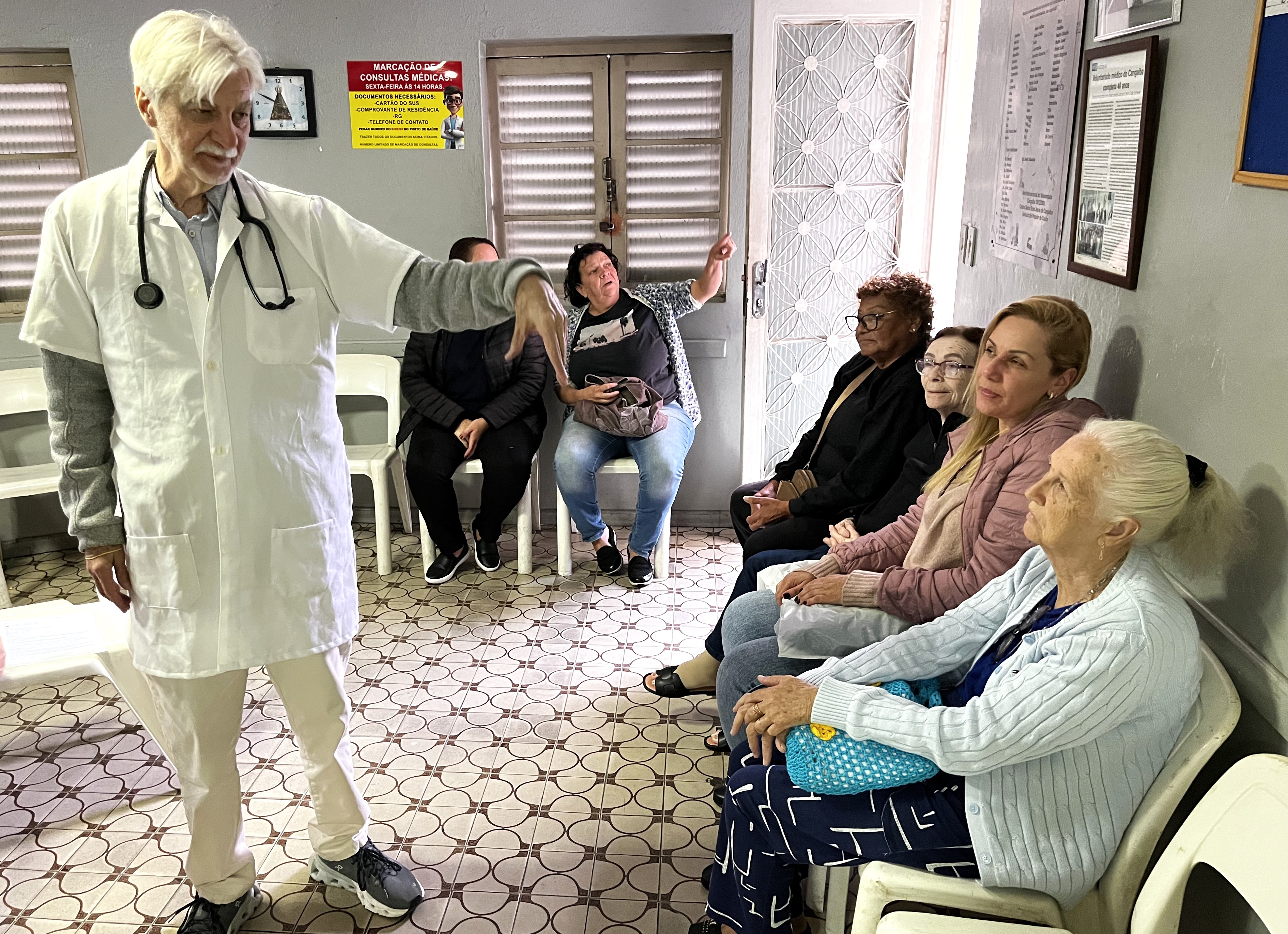 A imagem mostra uma atividade de educação em saúde realizada em uma sala de espera de uma unidade de saúde. Em primeiro plano, um profissional de saúde, vestindo jaleco branco e com um estetoscópio pendurado no pescoço, conversa com um grupo de pacientes. Ele faz um gesto com a mão enquanto parece explicar algum tema ou orientar os presentes. Sentadas em cadeiras ao longo da parede, estão principalmente mulheres, incluindo pessoas idosas, que acompanham atentamente a atividade. O ambiente é simples e acolhedor, com cartazes informativos, quadro de avisos, relógio e sinalizações relacionadas ao funcionamento do serviço de saúde. A cena transmite a ideia de cuidado próximo e humanizado, destacando o papel da Atenção Primária na promoção da saúde, na orientação dos usuários e na construção de vínculos entre profissionais e comunidade. A presença de pessoas idosas sugere que a atividade pode estar relacionada a temas como envelhecimento saudável, prevenção de doenças, autocuidado ou acompanhamento de condições crônicas.