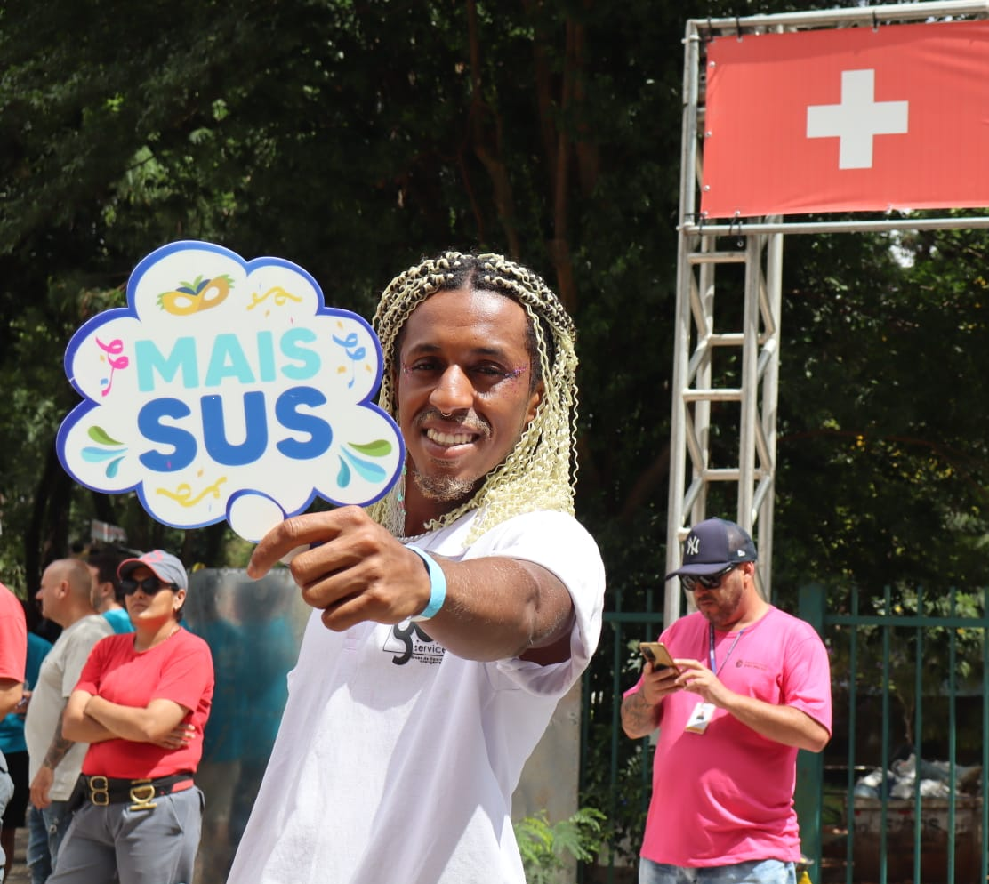 A imagem mostra um homem em primeiro plano sorrindo enquanto segura uma placa colorida em formato de balão com a frase “MAIS SUS” em uma rua. Ele veste uma camiseta branca e tem cabelos trançados claros. Ao fundo, há outras pessoas presentes. À direita, há uma estrutura metálica com uma bandeira vermelha que tem uma cruz branca no centro. 
