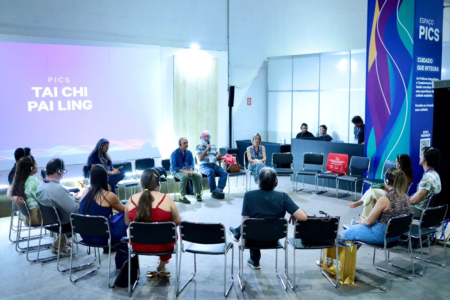 A imagem mostra um grupo de pessoas reunidas em um espaço fechado, organizado em formato de círculo com cadeiras. Parece ser uma roda de conversa ou atividade coletiva. No centro, há alguns participantes sentados, incluindo um homem mais velho com barba branca segurando um microfone, falando com o grupo. Ao lado dele, outras pessoas também estão sentadas, ouvindo atentamente. Todos os participantes ao redor usam fones de ouvido. Ao fundo, há uma projeção em uma tela com o texto “PICS TAI CHI PAI LING”, indicando que o encontro está relacionado a práticas integrativas de saúde, possivelmente envolvendo Tai Chi. À direita, há um painel vertical colorido com o título “ESPAÇO PICS – Cuidado que integra”, reforçando o tema do evento. O ambiente é de um congresso de saúde, com iluminação forte e estrutura de estandes. A atmosfera é tranquila e focada, com as pessoas participando de forma atenta e organizada.