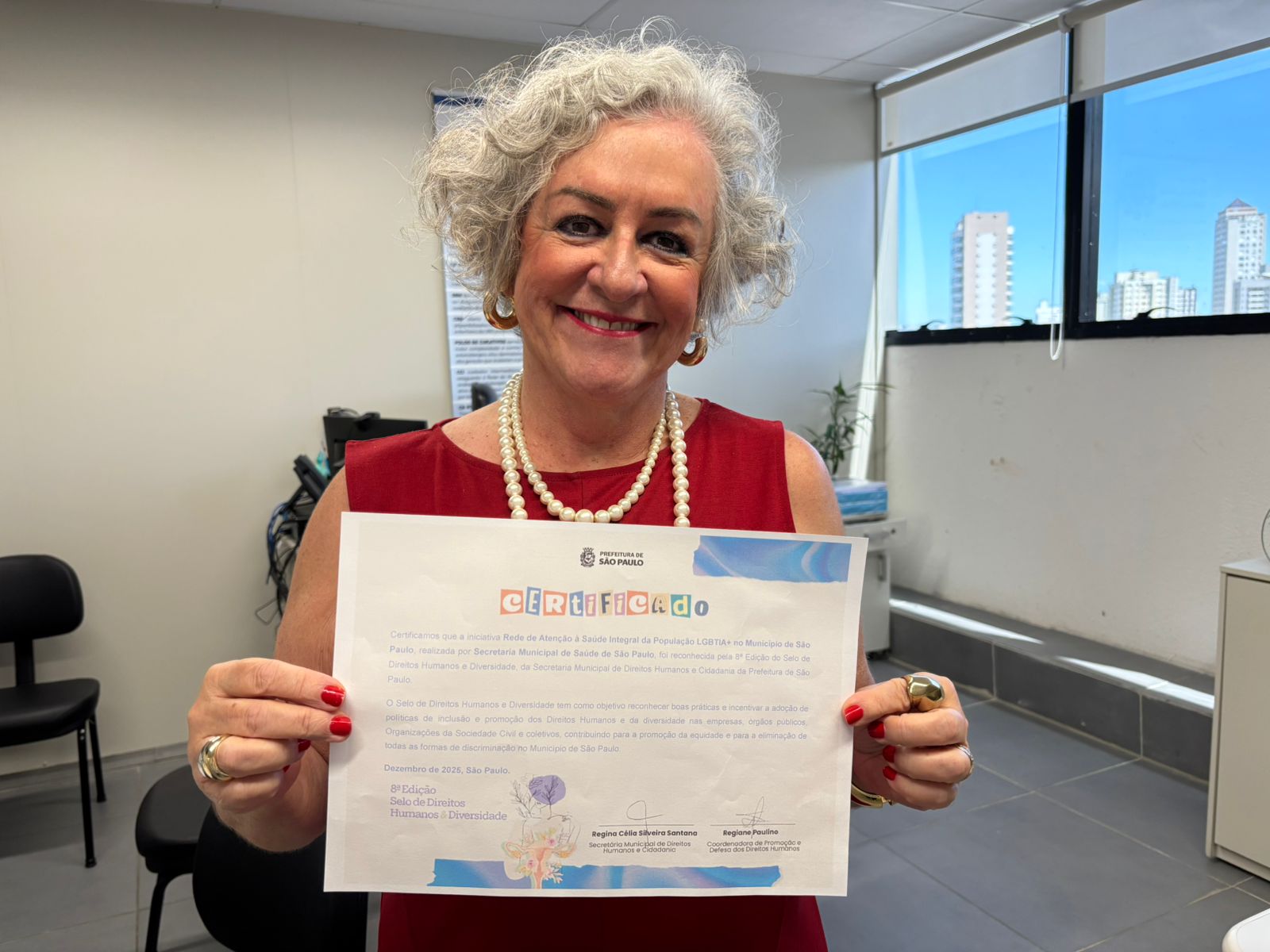 A imagem mostra uma mulher sorridente, com cabelo curto e grisalho, usa brincos dourados, um colar de pérolas e um vestido vermelho. Ela segura o certificado de premiação da Rede SAMPA Trans na 8ª edição do Selo de Direitos Humanos e Diversidade da Prefeitura de São Paulo.