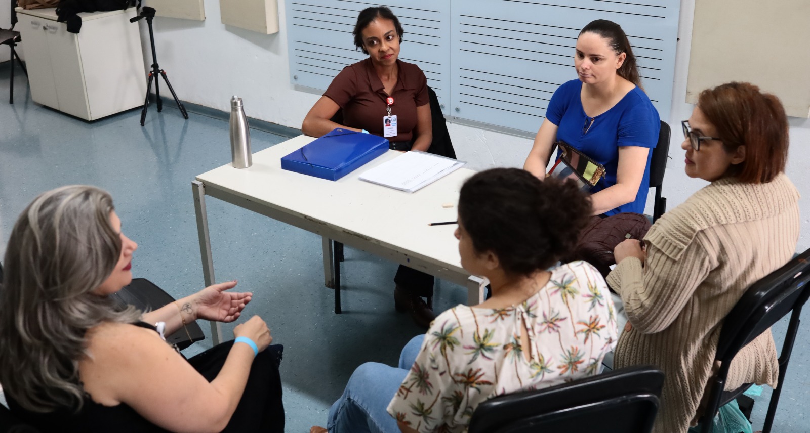 A imagem mostra um pequeno grupo de pessoas reunidas em uma sala que parece ser de um serviço público, possivelmente uma unidade de saúde ou espaço de atendimento. Cinco mulheres estão sentadas ao redor de uma mesa simples. Duas delas, posicionadas atrás da mesa, parecem conduzir a conversa — uma usa crachá e tem uma pasta azul à frente, sugerindo que pode ser uma profissional de saúde ou facilitadora. As outras três mulheres estão sentadas do lado oposto, ouvindo com atenção; uma delas gesticula enquanto fala, indicando que está compartilhando alguma experiência ou fazendo uma pergunta. Sobre a mesa há alguns materiais de trabalho, como uma pasta, papéis e uma garrafa térmica.