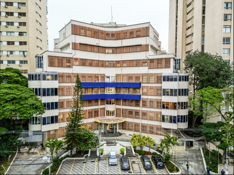A imagem mostra a fachada de um grande edifício hospitalar urbano, de vários pavimentos, com arquitetura simétrica e formato levemente escalonado. O prédio é predominantemente claro, em tons de bege e branco, com longas fileiras de janelas protegidas por brises verticais de cor amadeirada, que percorrem quase toda a extensão das fachadas.  No centro do edifício, há um volume destacado com uma faixa horizontal azul, marcando a área principal de acesso. A entrada fica no nível térreo, com escadarias e uma marquise curva, onde é possível identificar a sinalização do hospital. À frente, observa-se uma área de estacionamento com vagas demarcadas e alguns carros estacionados.  O entorno é composto por árvores de diferentes portes, incluindo palmeiras e uma árvore alta e estreita próxima ao eixo central do prédio, que suavizam a paisagem urbana. Ao fundo e nas laterais, aparecem edifícios residenciais altos, reforçando o contexto de uma região densamente urbanizada. A foto foi feita em perspectiva frontal e elevada, permitindo uma visão ampla da fachada e do espaço externo do hospital.