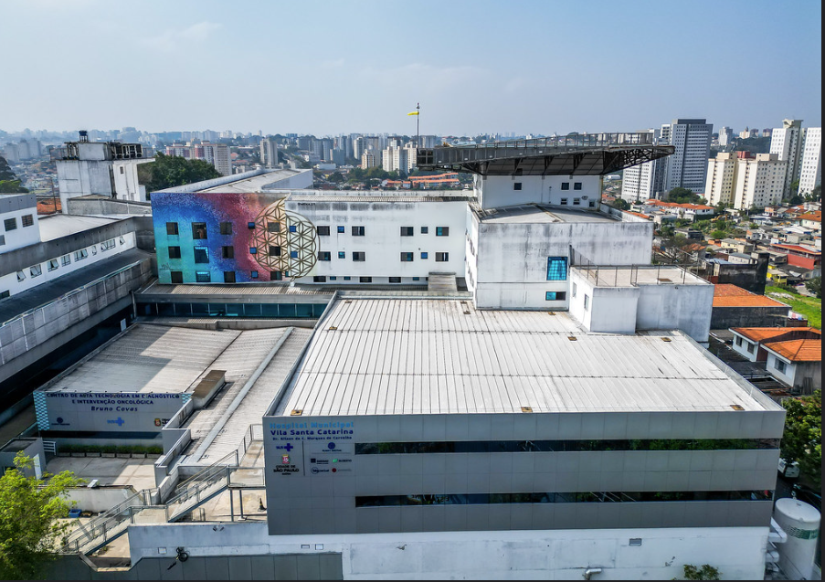 A imagem mostra uma vista aérea do Hospital Municipal Gilson de Cássia Marques de Carvalho – Vila Santa Catarina, evidenciando sua estrutura ampla e integrada ao tecido urbano da Zona Sul de São Paulo.  O complexo hospitalar é formado por edifícios de grande porte, predominantemente em tons claros, com volumes geométricos bem definidos e cobertura plana. Em destaque, na lateral de um dos prédios, há um grande mural artístico colorido, que traz formas geométricas e um rosto estilizado, conferindo identidade visual e humanização ao espaço hospitalar.  Na fachada frontal, é possível identificar a identificação institucional do hospital, com a logomarca da Prefeitura de São Paulo e do Sistema Único de Saúde (SUS). Também aparece a sinalização do Centro de Alta Tecnologia em Diagnóstico e Intervenção Oncológica Bruno Covas, reforçando a vocação da unidade para o atendimento oncológico de alta complexidade.