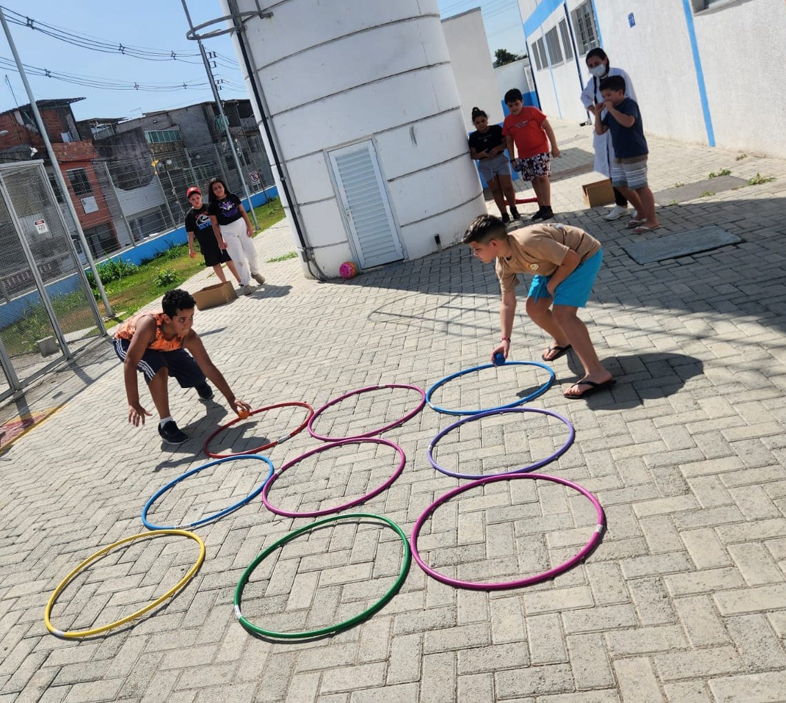 A imagem mostra uma atividade recreativa ao ar livre realizada em um pátio pavimentado. No centro da cena, vários bambolês coloridos estão dispostos no chão em fileiras, formando um percurso ou área de jogo.  Em primeiro plano, duas crianças participam da atividade. Cada uma está inclinada para frente, concentrada, segurando pequenas bolas coloridas e posicionando-as dentro dos bambolês. A postura dos participantes sugere uma dinâmica de competição ou cooperação que exige coordenação motora e atenção.  Ao fundo, outras crianças observam a brincadeira enquanto aguardam sua vez. Também há uma profissional de saúde vestindo jaleco branco e máscara, acompanhando a atividade. O ambiente parece ser uma área externa de uma instituição, com prédios de paredes brancas e detalhes em azul, além de um muro, cercas e construções residenciais visíveis ao redor.  A cena é iluminada pela luz do dia e transmite um clima de interação, lazer e promoção de atividades físicas e coletivas para crianças.