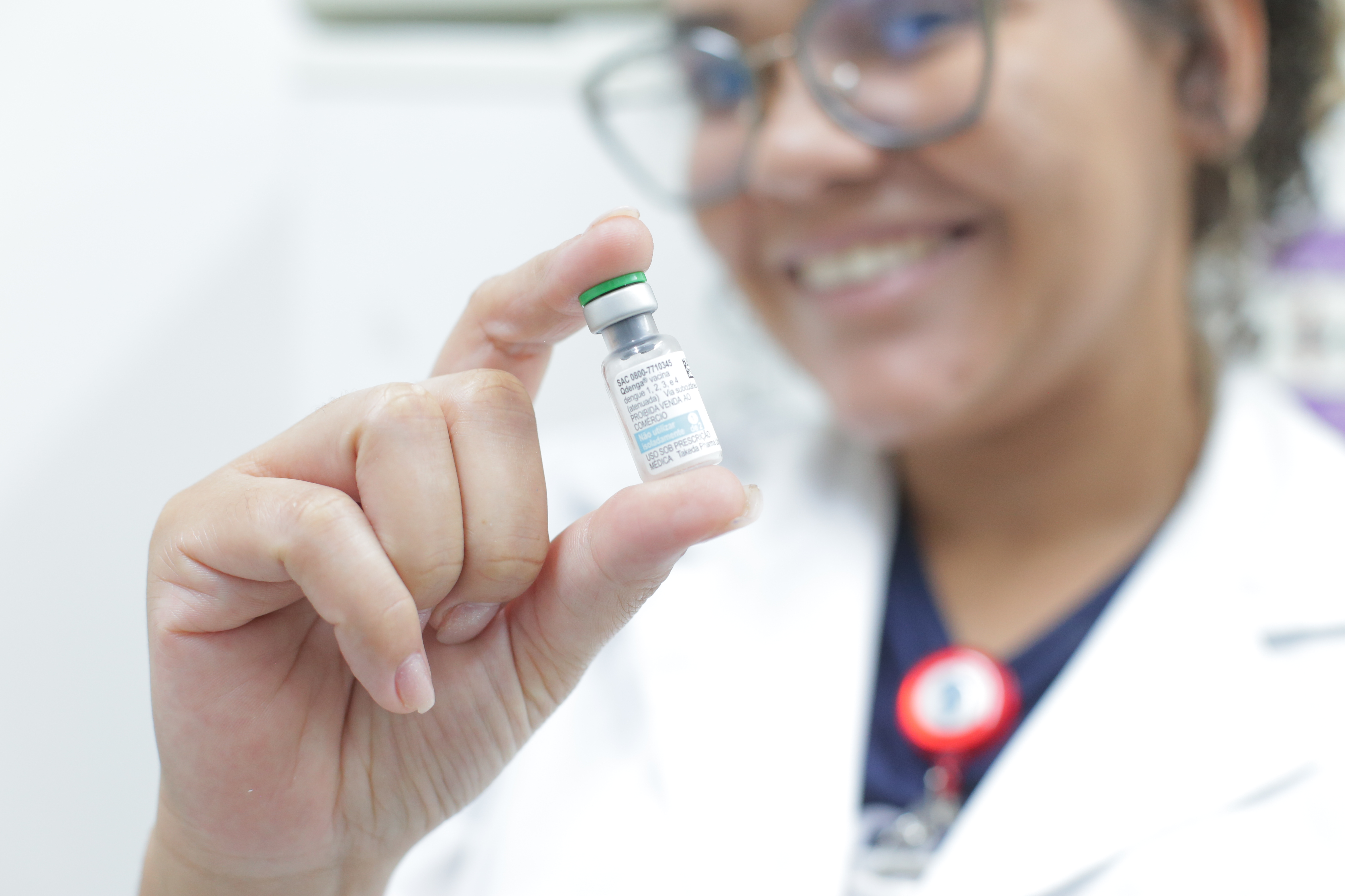 A imagem mostra uma profissional de saúde sorrindo enquanto segura um frasco de vacina entre os dedos, em destaque no primeiro plano. Ela veste jaleco branco e crachá, e usa óculos de grau. O foco da foto está no pequeno frasco com tampa verde, identificado por um rótulo, enquanto o rosto da profissional aparece levemente desfocado ao fundo. O ambiente é claro, típico de uma unidade de saúde, transmitindo uma sensação de cuidado e confiança.