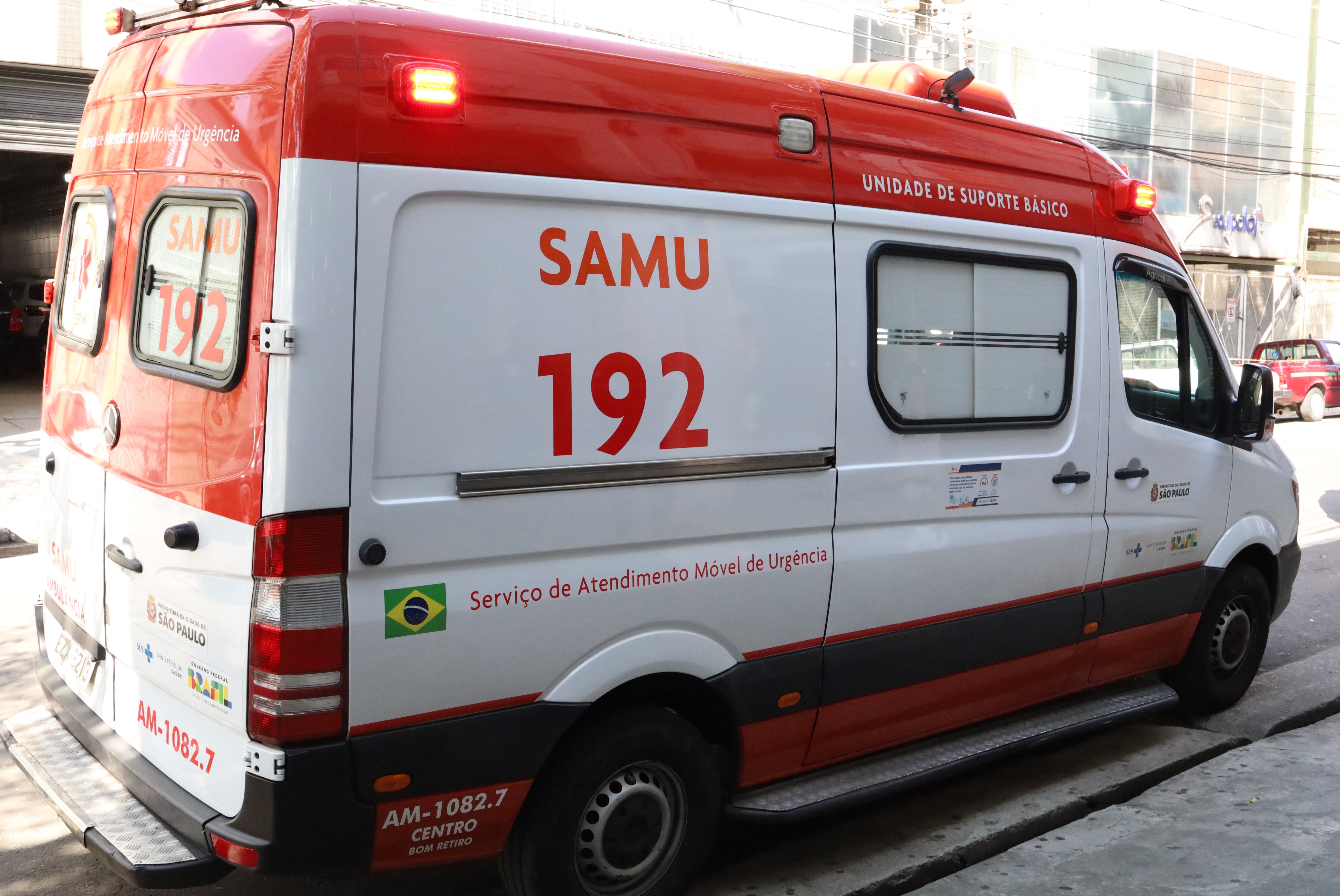 A imagem mostra uma ambulância do SAMU (Serviço de Atendimento Móvel de Urgência), estacionada em uma rua. O veículo é branco com detalhes em vermelho, tem a inscrição “SAMU 192” bem visível na lateral, número que corresponde ao telefone de emergência do serviço. Na lateral também estão escritos “Unidade de Suporte Básico” e “Serviço de Atendimento Móvel de Urgência”, junto com o logotipo da Prefeitura de São Paulo, o brasão do Brasil e a bandeira nacional. Na traseira, observa-se a identificação da unidade: AM-1082.7 – Centro / Bom Retiro. A ambulância é equipada com sirene e giroflex vermelhos, que estão acesos. Trata-se de uma viatura utilizada para atendimento pré-hospitalar e transporte de pacientes em situações de urgência.
