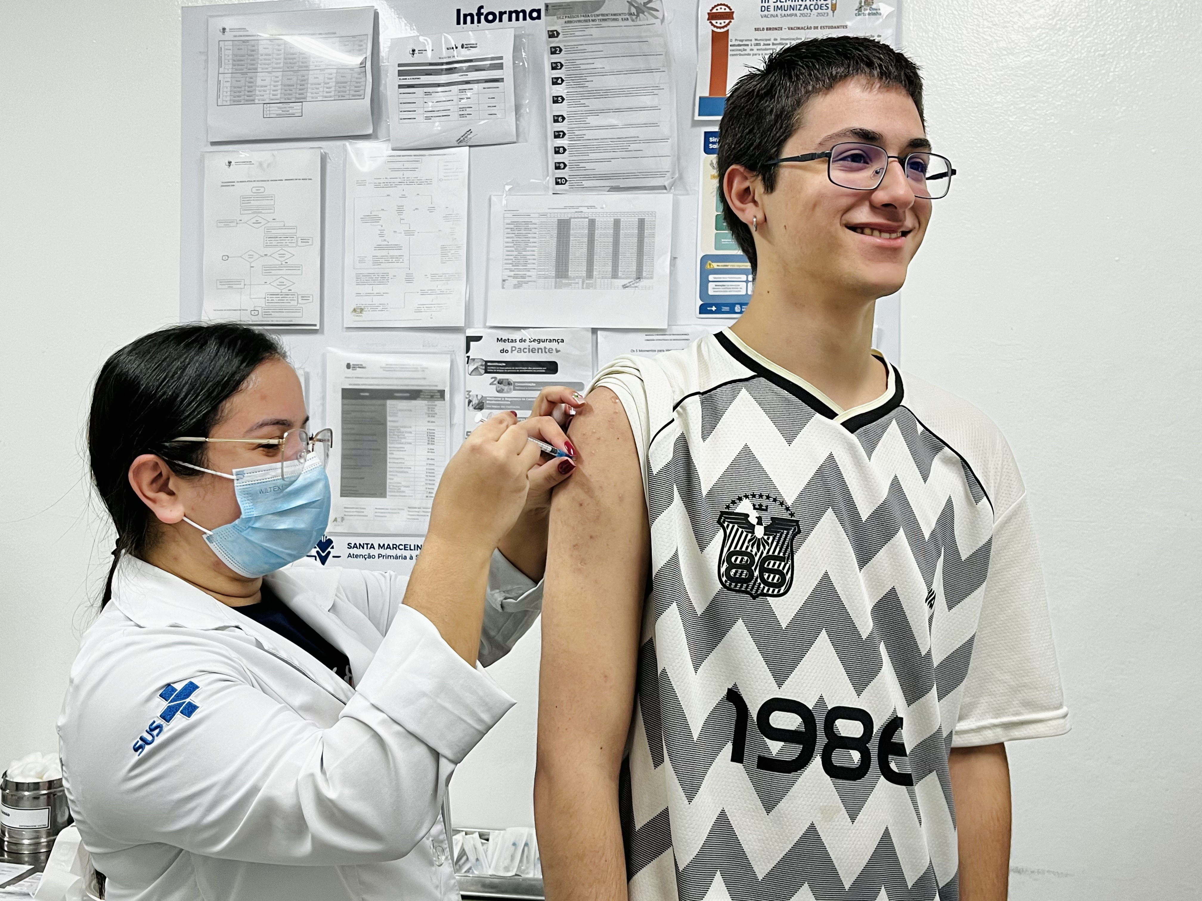 A imagem mostra uma profissional de saúde do SUS aplicando uma vacina no braço de um adolescente, em uma unidade de saúde. A profissional usa jaleco branco, máscara cirúrgica e óculos de proteção e segura uma seringa enquanto realiza a aplicação. O adolescente está em pé, sorrindo e olhando para fora do enquadramento. Ele veste uma camisa esportiva branca e preta. Ao fundo, há um painel com cartazes, tabelas, fluxogramas e orientações de saúde, incluindo materiais relacionados à imunização e à segurança do paciente. Também é possível identificar a referência à Santa Marcelina – Atenção Primária à Saúde.