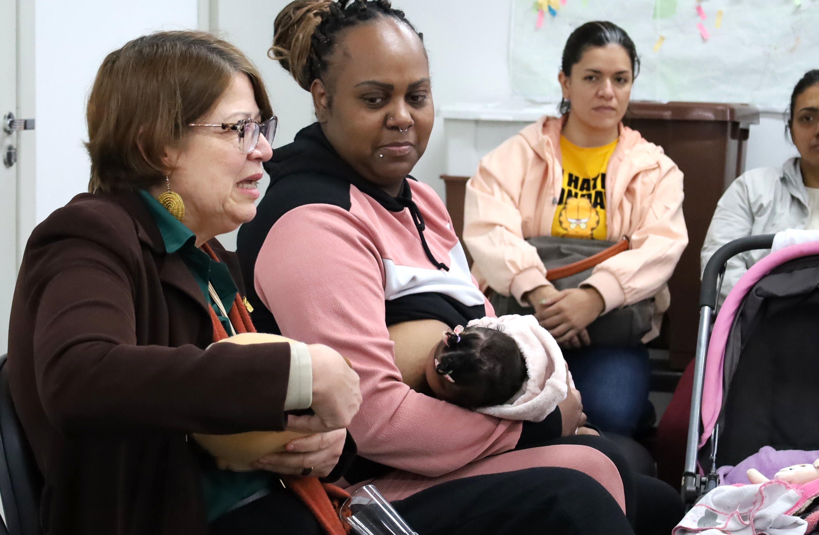 A imagem mostra um grupo de quatro mulheres reunidas em uma sala, aparentemente durante uma atividade ou encontro de caráter comunitário ou de saúde. Em primeiro plano, uma mulher de óculos, usando blazer escuro, camisa verde e brincos grandes, fala enquanto segura uma pequena tigela ou recipiente. Ao lado dela, uma mulher negra, vestindo conjunto esportivo rosa, preto e branco, segura no colo um bebê que está enrolado em uma manta clara. Ao fundo, outras duas mulheres acompanham a conversa; uma delas usa uma jaqueta clara sobre uma camiseta amarela, enquanto a outra aparece parcialmente à direita, ao lado de um carrinho de bebê. A cena transmite um momento de conversa, orientação ou troca de experiências em um ambiente institucional.  