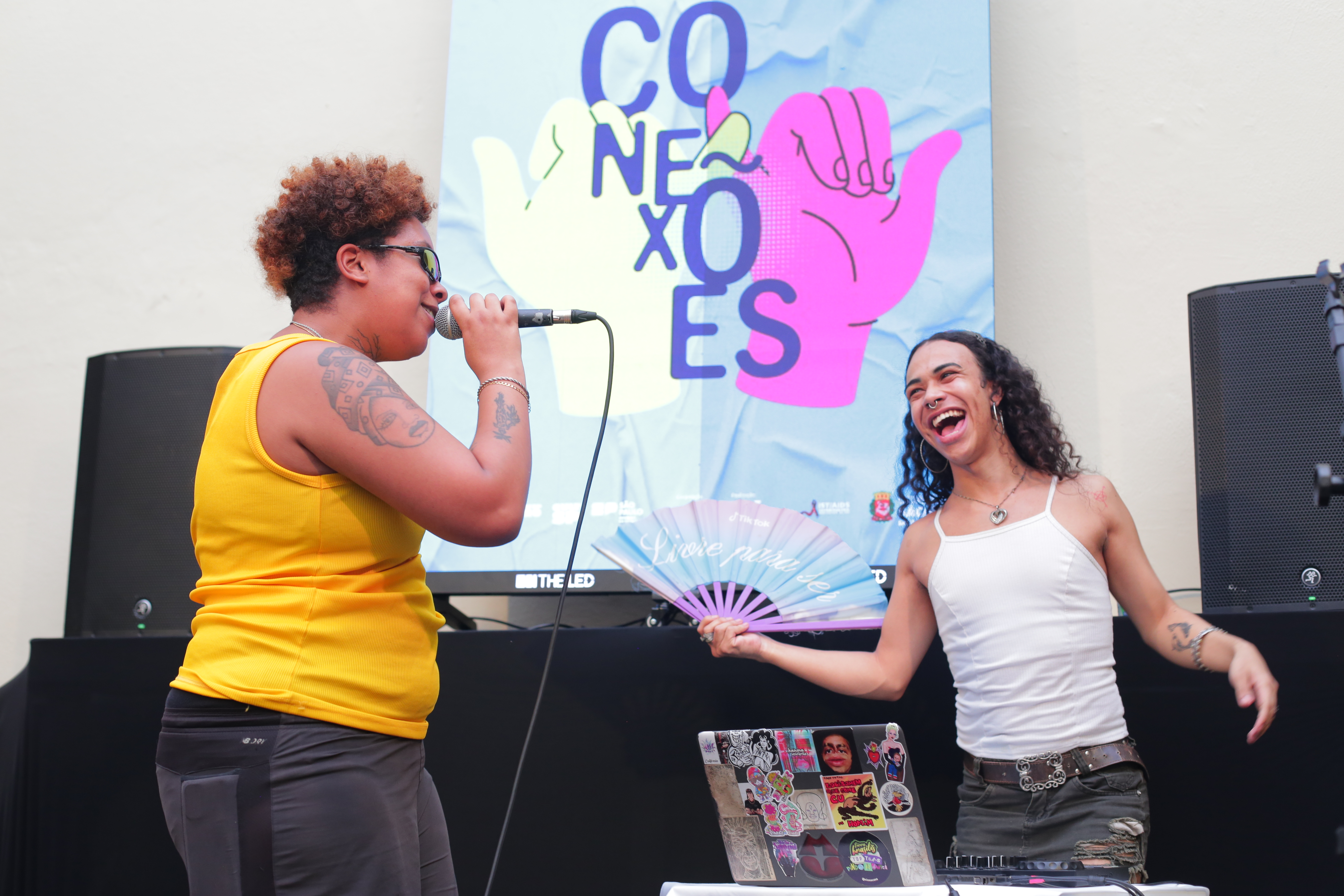 Apresentação de rap do Travas da Sul. À esquerda, uma mulher de cabelo curto cacheado e óculos de sol segura um microfone, vestindo uma blusa amarela e calça cinza. Ela tem tatuagens visíveis nos braços e está cantando. À direita, outra mulher sorri de forma animada, com cabelos longos e cacheados, vestindo uma regata branca e um cinto estiloso com uma saia ou shorts. Ela segura um leque colorido e está fazendo a discotecagem. Ao fundo, há um painel colorido com a palavra