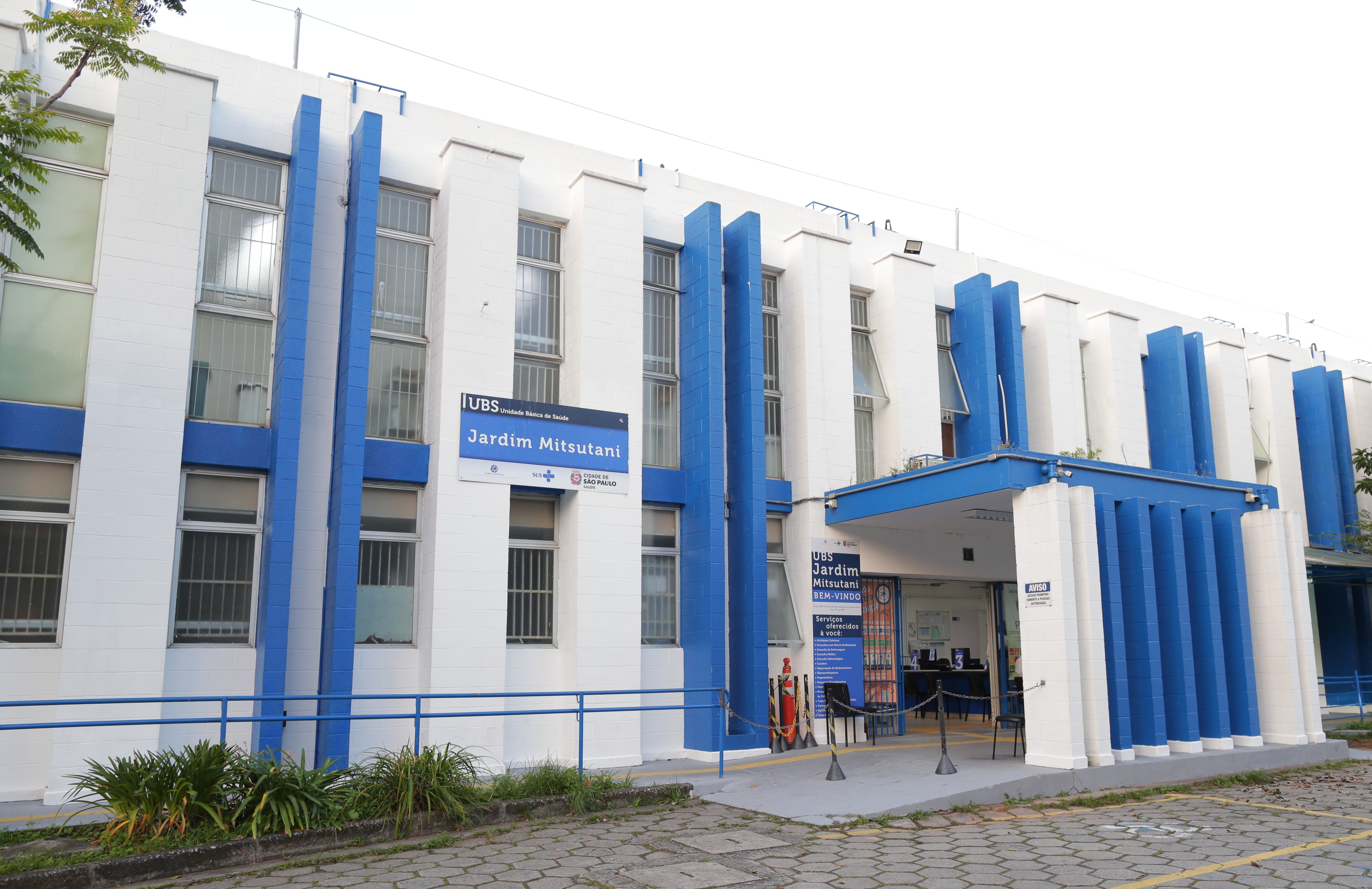 A imagem mostra a fachada de uma Unidade Básica de Saúde identificada como UBS Jardim Mitsutani. O prédio é de dois pavimentos, com arquitetura funcional e cores predominantes azul e branca, características comuns de equipamentos públicos de saúde. Na parte frontal, há uma marquise que cobre a entrada principal, com sinalização visível indicando o nome da unidade e mensagens de boas-vindas. As janelas são amplas, com grades, e o edifício possui rampas e corrimãos, evidenciando preocupação com acessibilidade. Em frente à UBS, observa-se uma área pavimentada para circulação de pedestres e acesso de usuários. O conjunto transmite a imagem de um equipamento público organizado, voltado ao atendimento da população local.