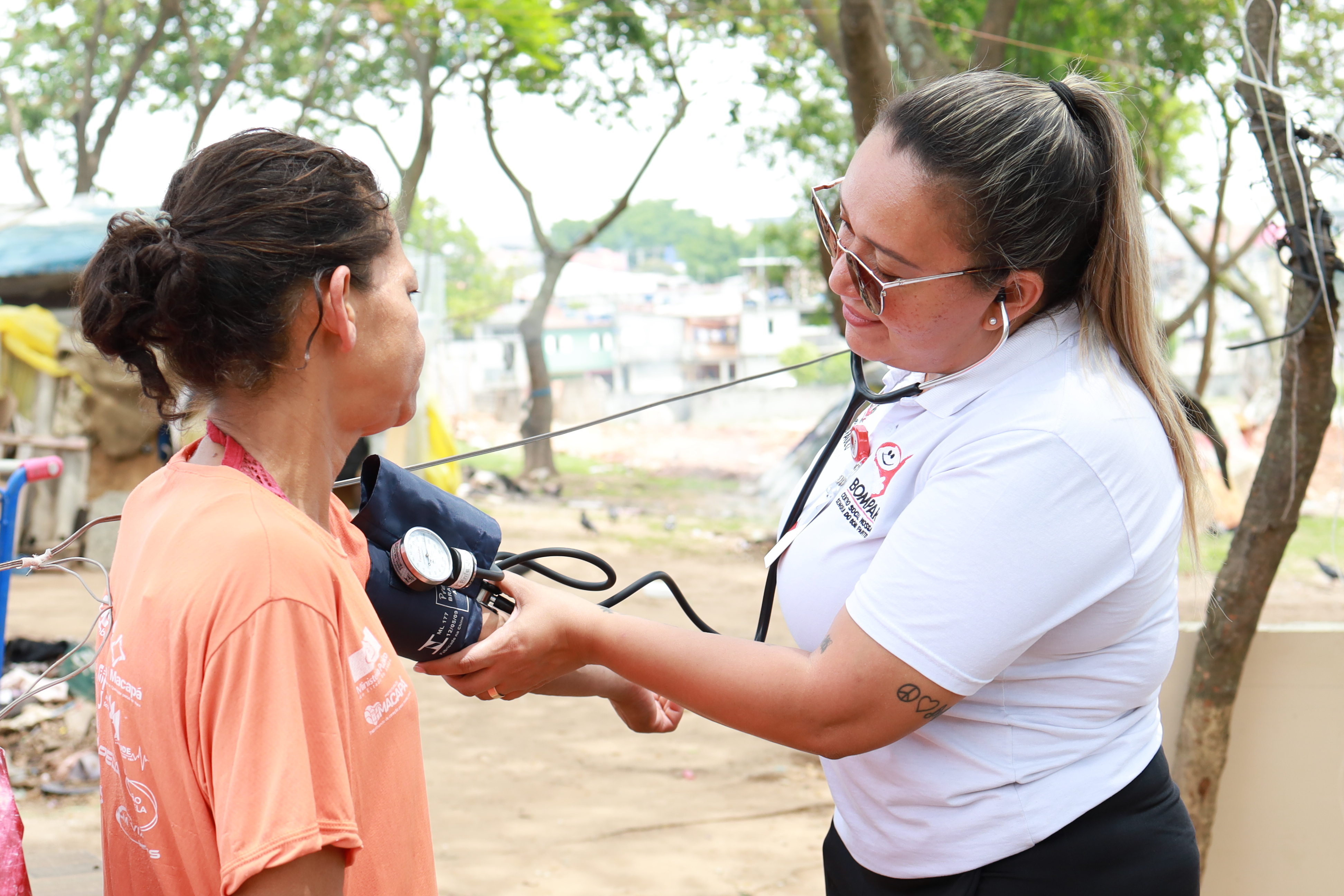 A imagem mostra uma profissional de saúde realizando a aferição da pressão arterial de uma mulher em um ambiente ao ar livre. A profissional, que veste uma camisa branca com logotipos de um projeto de assistência, utiliza um estetoscópio e um esfigmomanômetro (aparelho de pressão) para examinar a paciente, que está vestida com uma camiseta laranja. A cena sugere um atendimento comunitário ou ação social de saúde, possivelmente em uma área de vulnerabilidade. Ao fundo, há árvores, construções e sinais de uma ocupação urbana informal. A interação transmite cuidado, empatia e atenção às necessidades básicas de saúde da população