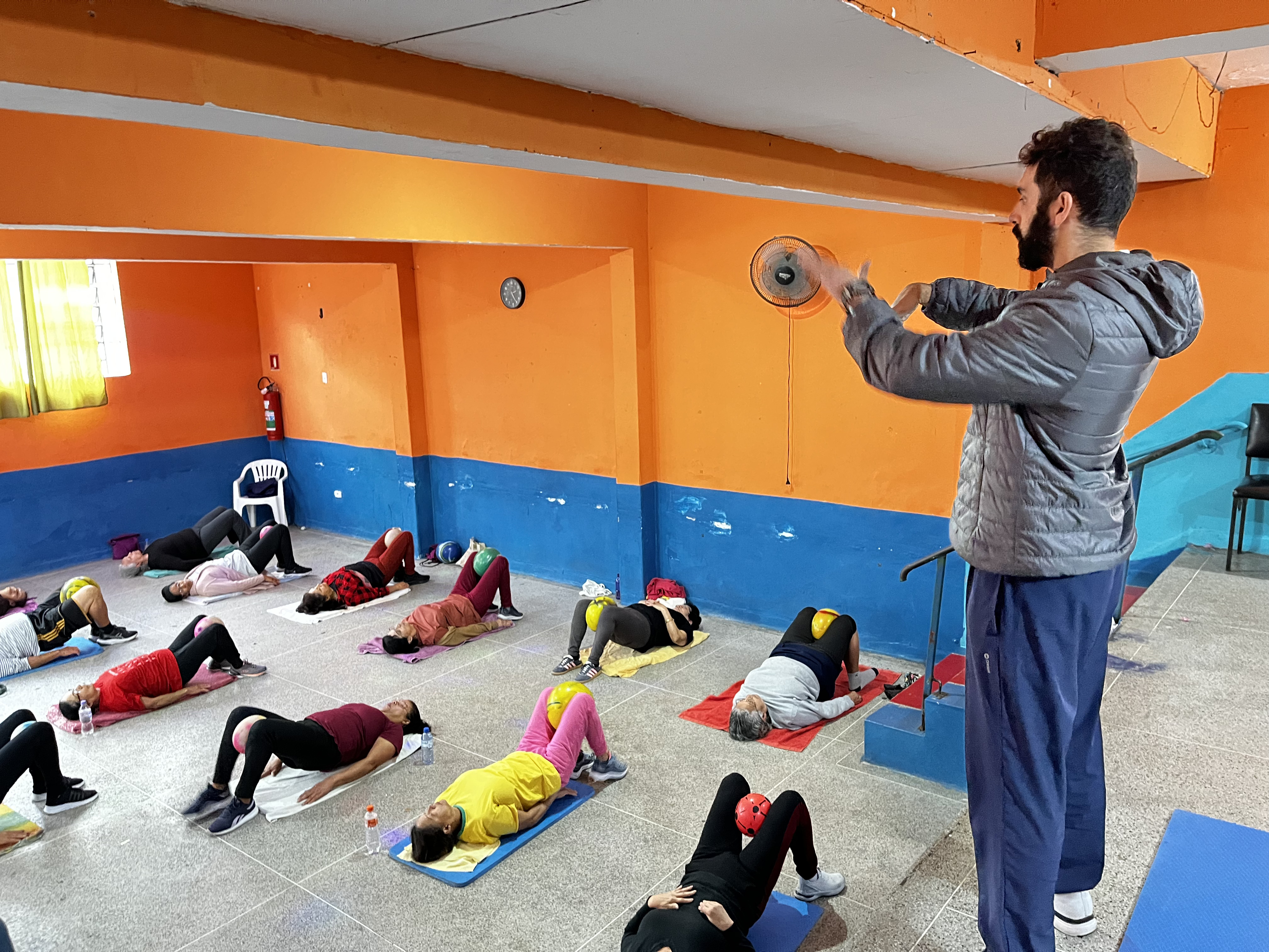 A imagem mostra uma atividade coletiva realizada em um espaço amplo, com paredes pintadas de laranja e azul. No chão, várias pessoas estão deitadas sobre colchonetes ou toalhas, realizando exercícios físicos orientados por um profissional que está à frente do grupo. Os participantes, em sua maioria adultos e idosos, fazem movimentos com as pernas e utilizam pequenas bolas coloridas posicionadas entre os joelhos ou sobre o corpo. O instrutor, vestido com roupa esportiva, demonstra um dos exercícios com as mãos. O ambiente conta ainda com ventiladores, corrimão e uma pequena área elevada, indicando um espaço adaptado para atividades físicas. A cena transmite a ideia de uma prática coletiva voltada ao movimento, à convivência e à promoção da saúde.