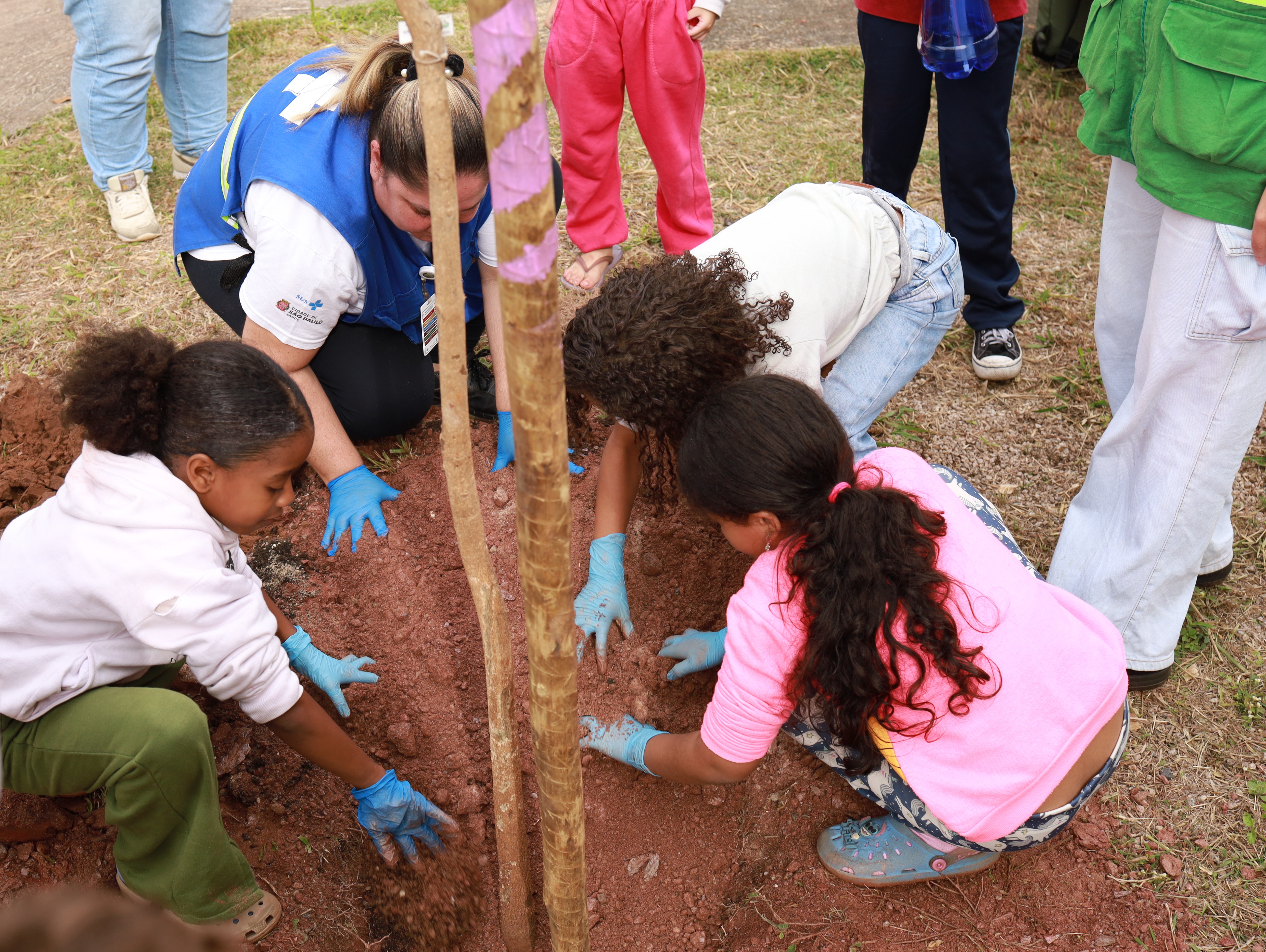 A imagem mostra um grupo de pessoas, incluindo uma adulta de uniforme e três meninas, agachadas em torno de uma muda de árvore, participando ativamente do plantio em um terreno gramado.