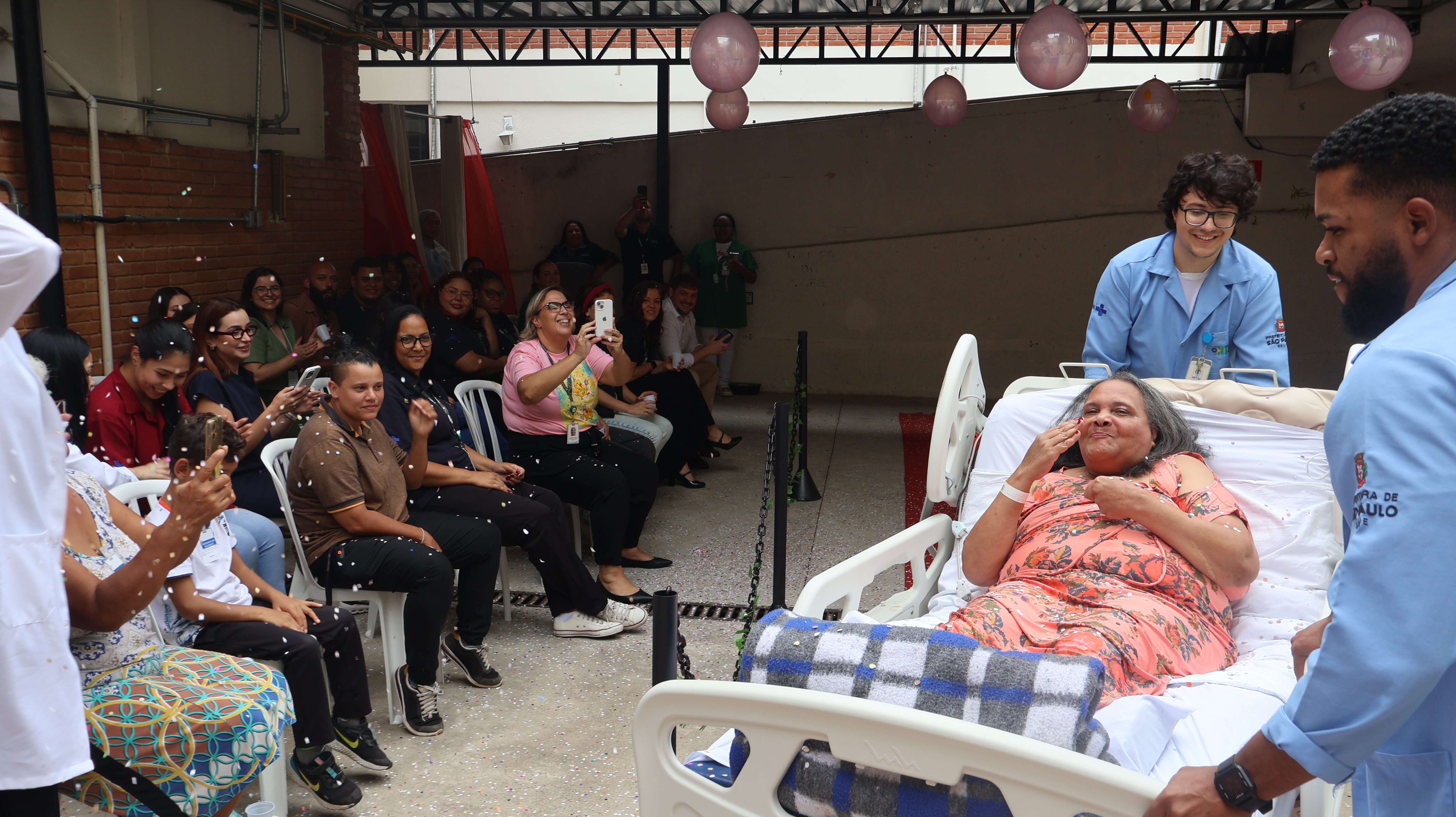 A fotografia registra um evento festivo em uma área externa ou pátio coberto do Hospital Municipal Guarapiranga. No centro da composição, uma paciente idosa está deitada em uma cama hospitalar branca, vestindo um robe floral rosa e coberta por uma manta xadrez azul. Ela sorri e faz um gesto de beijo para a plateia, visivelmente emocionada. A cama é conduzida por dois profissionais de saúde vestindo jalecos azuis claros com o logotipo da Prefeitura de São Paulo. Ao lado esquerdo, uma plateia formada por funcionários e familiares está sentada em cadeiras brancas, aplaudindo, sorrindo e registrando o momento com celulares. O ambiente está decorado com balões cor-de-rosa no teto e confetes brancos caem sobre a paciente, criando uma atmosfera de desfile e celebração.