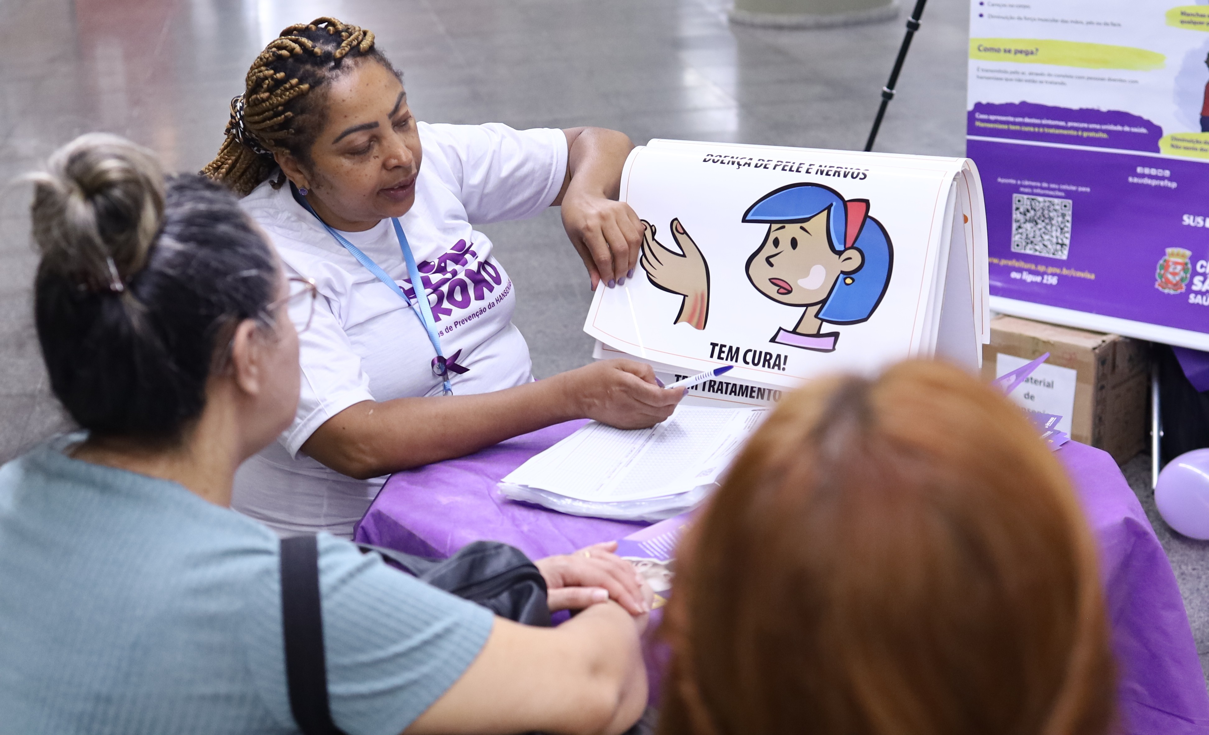 A imagem mostra uma ação educativa em saúde realizada em um espaço público ou institucional. No centro da cena, uma mulher, aparentemente profissional da saúde ou educadora, sentada atrás de uma mesa coberta com um tecido roxo, orienta outras mulheres. Ela veste uma camiseta branca com estampa roxa relacionada a uma campanha de saúde e usa um crachá no pescoço.  A profissional segura um material ilustrado em formato de cartaz ou flip chart, com desenhos de um rosto e uma mão, acompanhado da frase “Tem cura!”, indicando uma abordagem didática e acessível. À sua frente, duas mulheres estão sentadas, vistas de costas ou de perfil, atentas à explicação.  Ao fundo, há banners informativos com cores roxa e amarela, além de logotipos institucionais, sugerindo uma campanha de conscientização — possivelmente ligada à saúde da mulher ou à prevenção de doenças. O ambiente é bem iluminado e organizado, reforçando o caráter educativo, acolhedor e informativo da atividade.