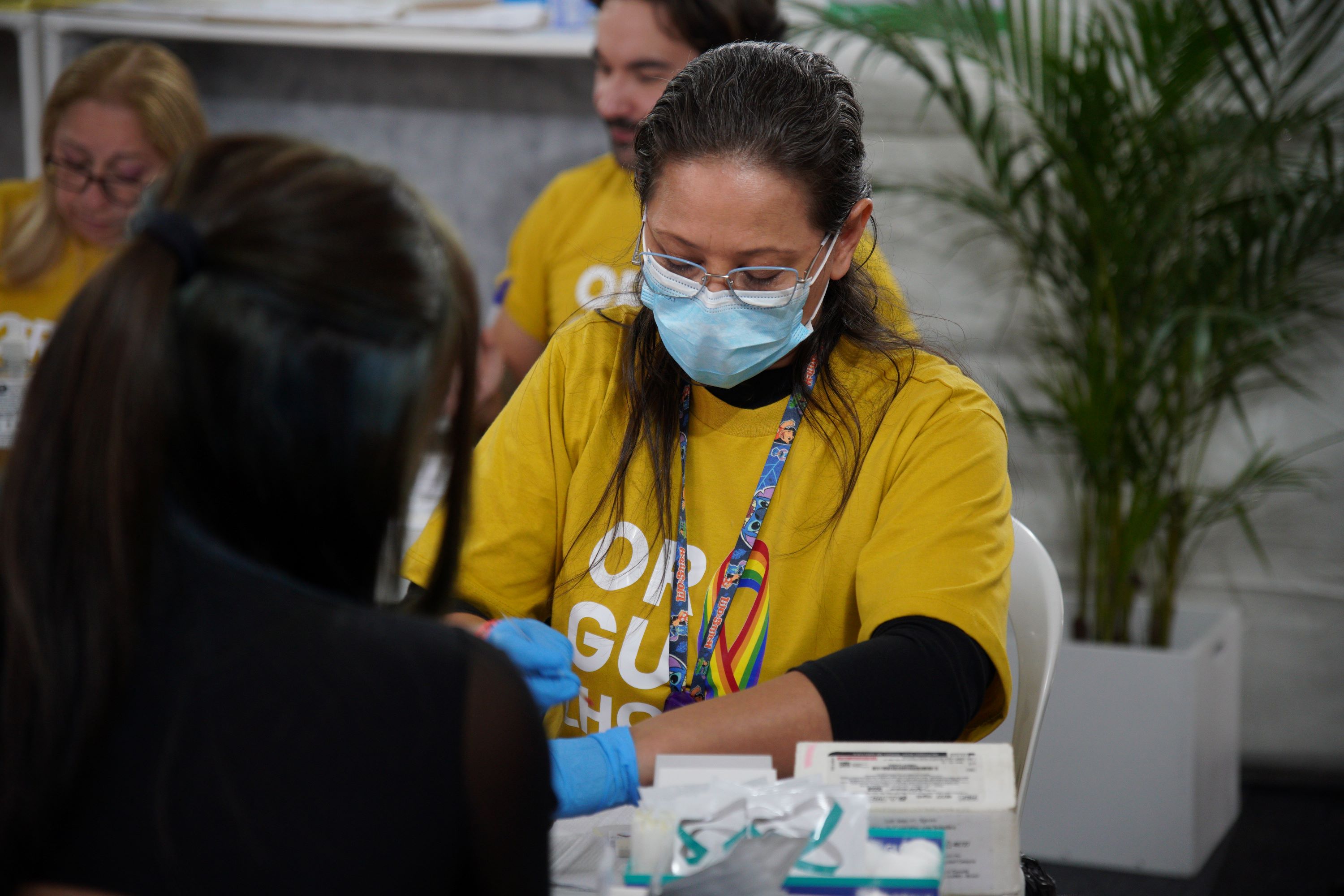 Profissional de saúde usando máscara, óculos e luvas azuis realiza atendimento a uma pessoa durante uma ação de prevenção em saúde. Ao fundo, outras pessoas da equipe, também com camisetas amarelas, participam da atividade.
