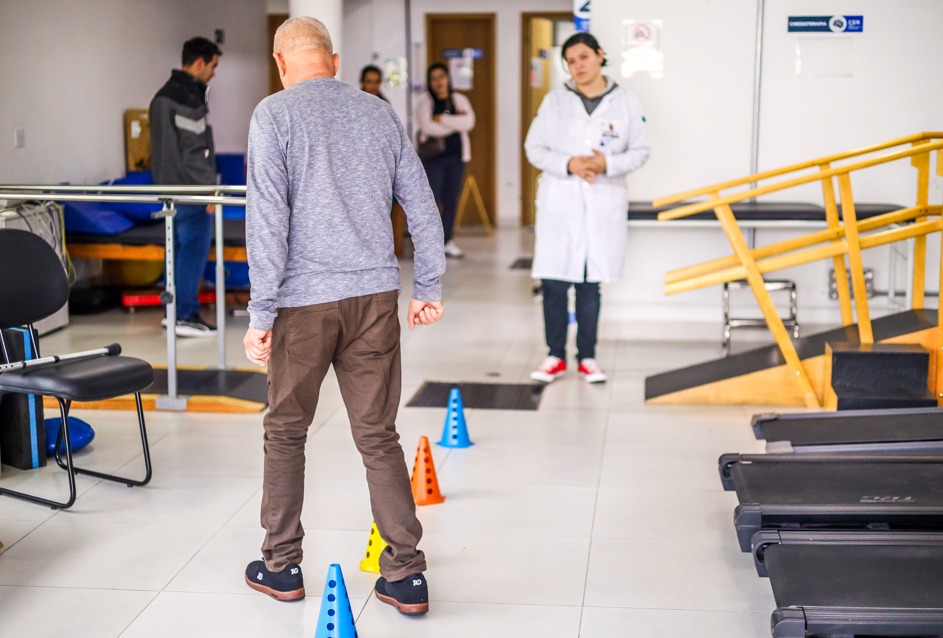 A imagem mostra um ambiente de fisioterapia. No centro, há um homem idoso, visto de costas, de pele clara e com cabelo curto e grisalho. Ele veste uma blusa de manga longa cinza, calça marrom e tênis pretos. O homem caminha entre cones coloridos (azul, amarelo e laranja) que estão posicionados no chão em linha, aparentemente como parte de um exercício de coordenação ou equilíbrio.  Ao fundo, há um profissional de saúde usando jaleco branco e tênis vermelhos, observando atentamente o exercício. Outras pessoas estão presentes no ambiente, algumas de pé, em segundo plano. À direita, vê-se um corrimão de madeira inclinado, típico para exercícios de fisioterapia, e esteiras de academia. À esquerda, há cadeiras e barras paralelas, também comuns em clínicas de reabilitação.  A cena transmite um momento de acompanhamento profissional em um treinamento físico voltado à recuperação da mobilidade.