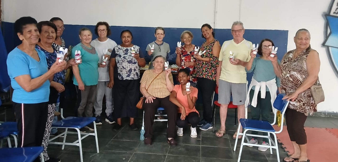 A foto mostra um grupo de aproximadamente 15 pessoas, a maioria idosas, reunidas em um espaço interno de paredes brancas e azul-marinho. Estão em pé, lado a lado, formando uma fileira, enquanto duas mulheres aparecem sentadas na frente, uma delas em uma cadeira de rodas. Todos parecem sorridentes e alegres.  Eles seguram pequenas lembrancinhas em formato de coelho branco, provavelmente feitas de papel ou tecido, com detalhes coloridos, sugerindo uma atividade de artesanato ou celebração, talvez ligada à Páscoa.  As pessoas vestem roupas casuais e confortáveis: camisetas, calças e blusas leves. Há diversidade nos estilos — desde estampas florais até roupas lisas. O ambiente tem cadeiras azuis de plástico e metal, algumas organizadas e outras fora de lugar, o que dá a sensação de um encontro descontraído e comunitário.